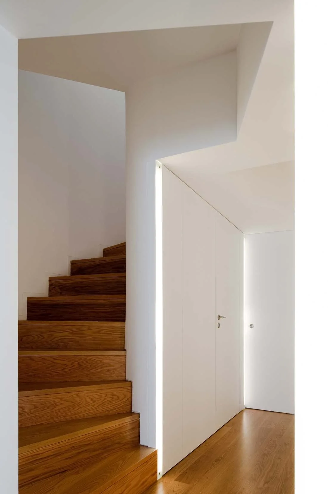 A minimalist hallway featuring a wooden staircase next to white walls with integrated vertical recessed strip lights and seamless white doors.