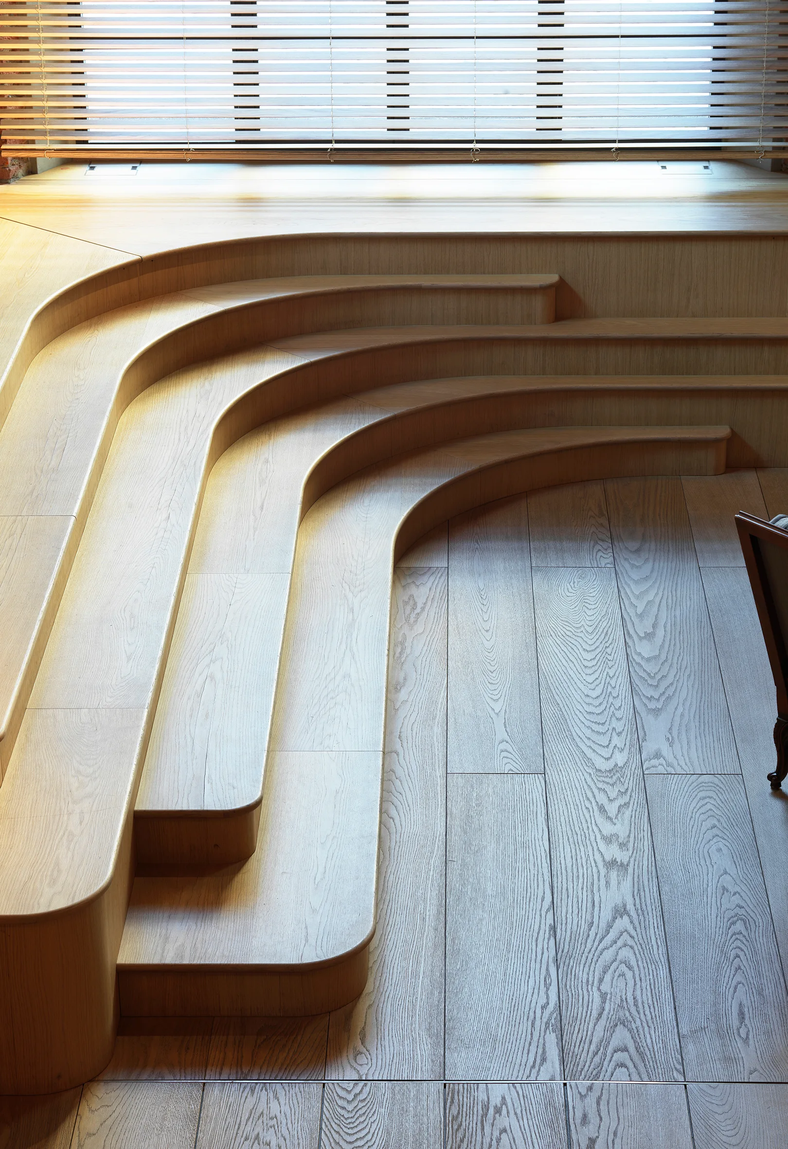 A custom curved light wood tiered platform with steps is situated below a window with horizontal blinds on a matching oak plank floor.