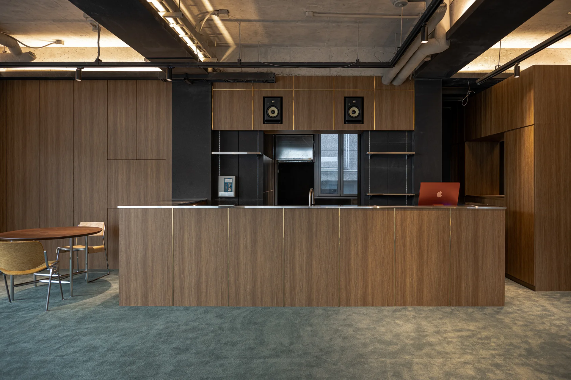 A modern kitchen area features a large wood-paneled island with metallic details, matching cabinetry, a teal carpeted floor, and an exposed concrete ceiling.