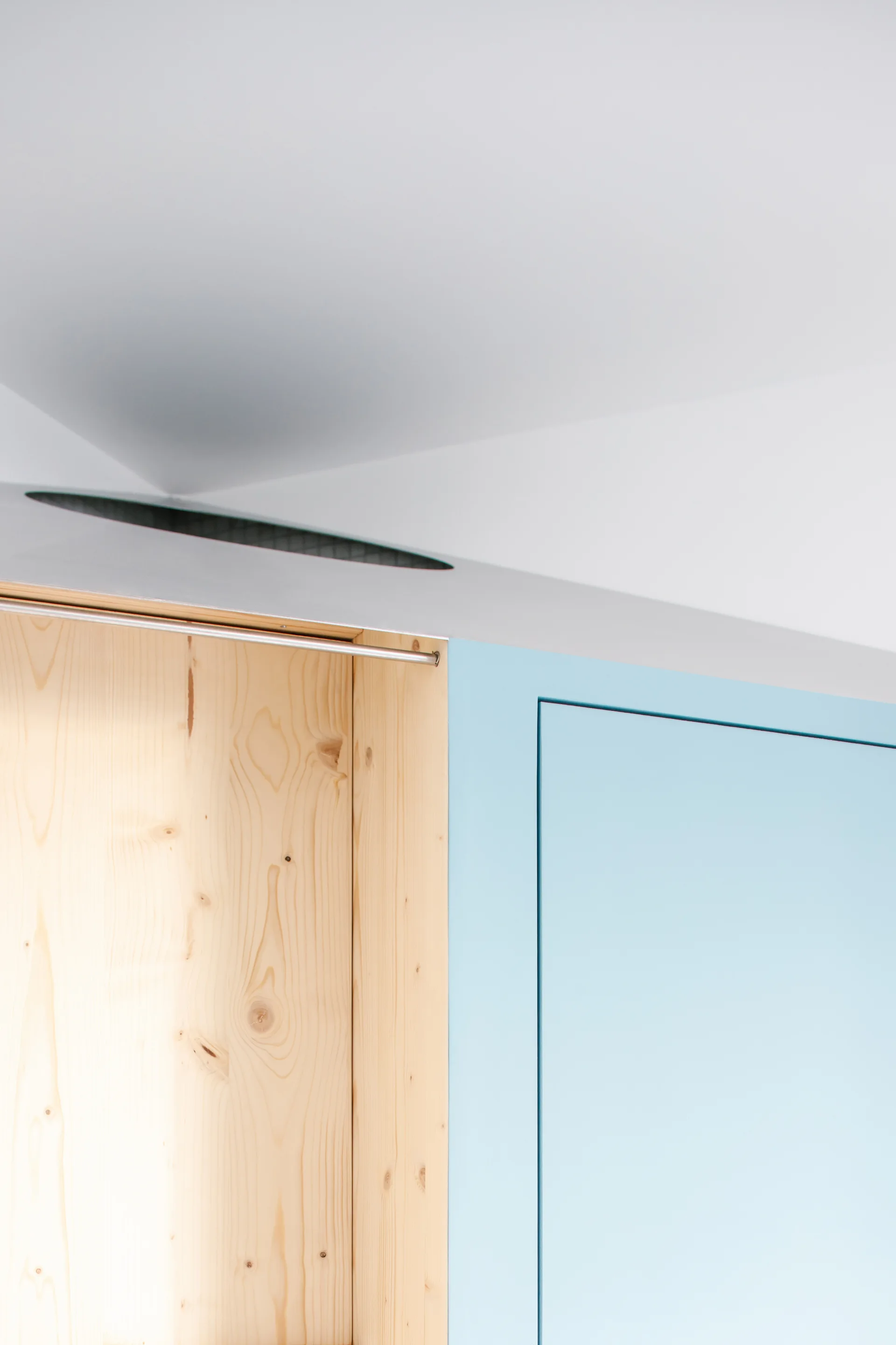 Detail of custom minimalist cabinetry featuring matte light blue wardrobe doors adjacent to a natural pine wood alcove with a slim metal rod.