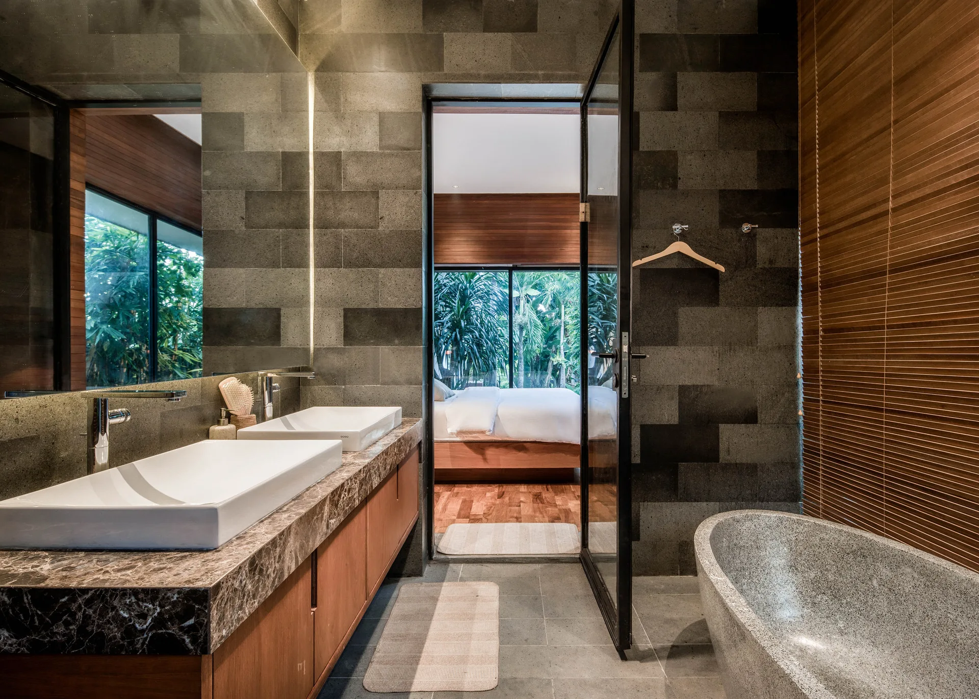 A modern ensuite bathroom features textured dark grey stone walls, a marble double vanity, a stone freestanding tub, and a glass door leading to a bedroom.