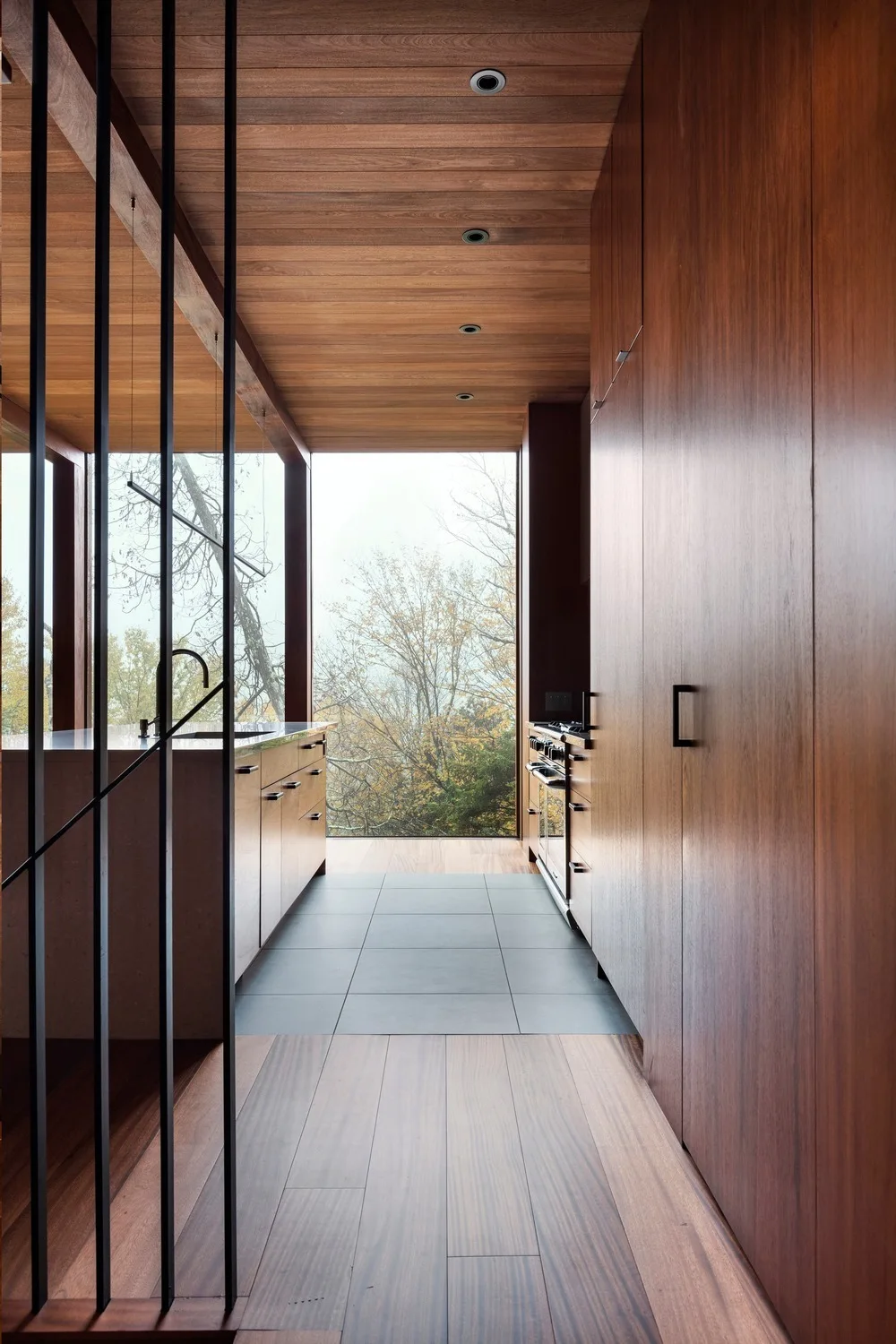 A modern galley kitchen featuring rich wood paneled walls, ceiling, and cabinets, a dark grey tiled floor runner, and a window framing trees.
