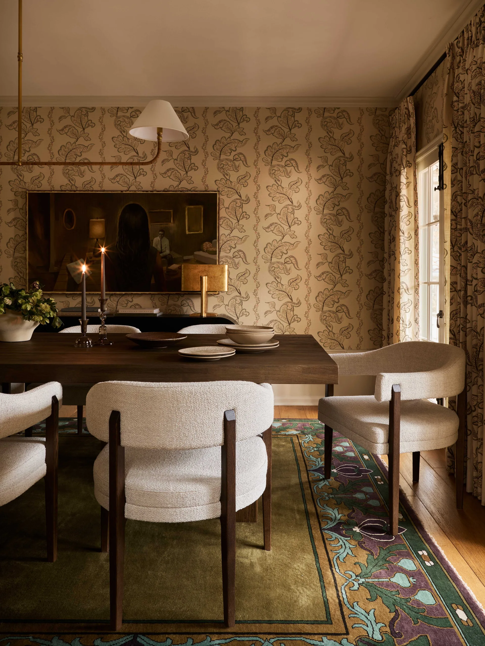 Textured cream dining chairs surround a dark wood table in a dining room with botanical patterned wallpaper, a green patterned rug, and a framed painting.