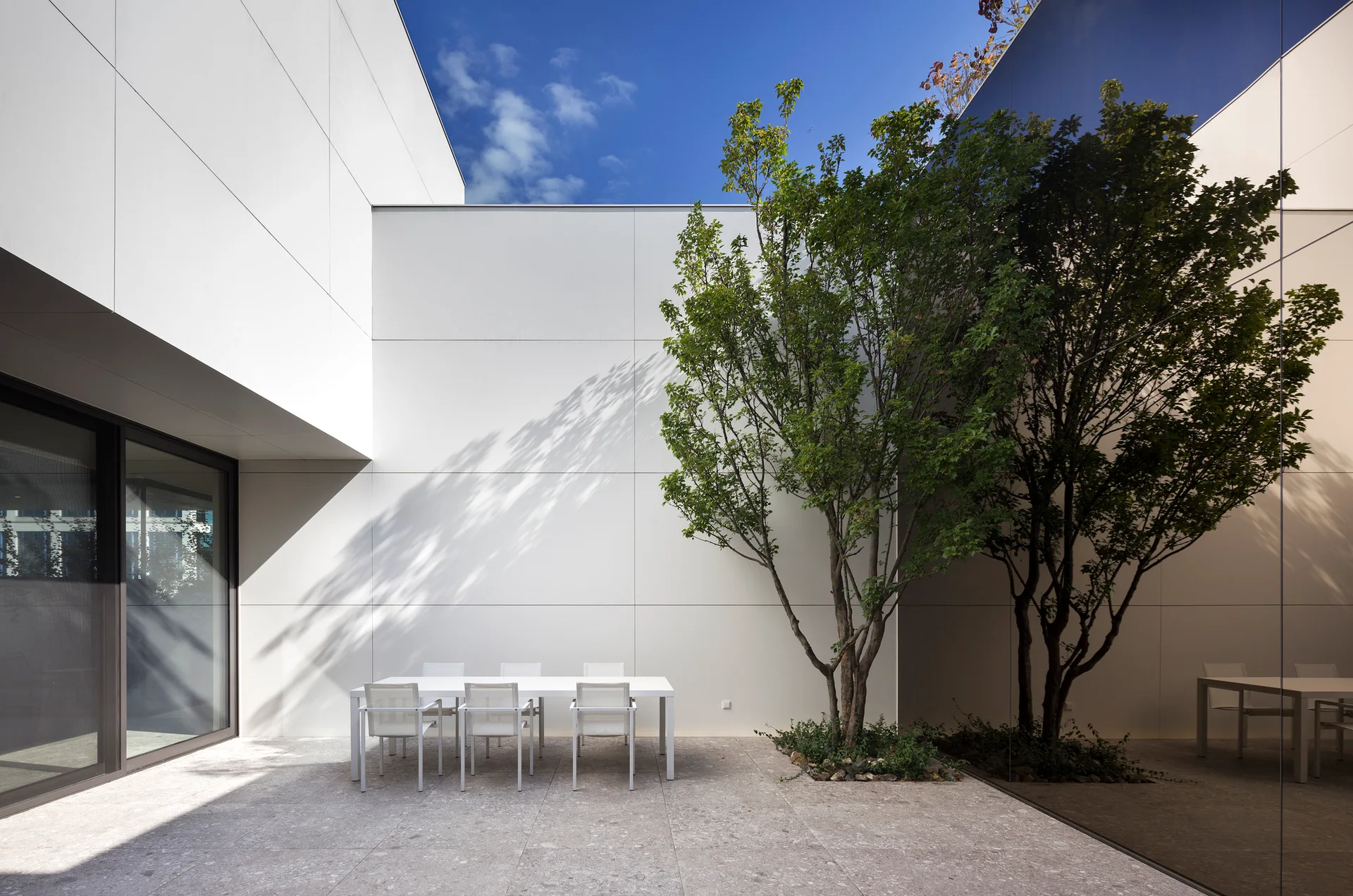 A minimalist outdoor patio features a white dining table and chairs on stone pavers next to a green tree and a large reflective glass wall.