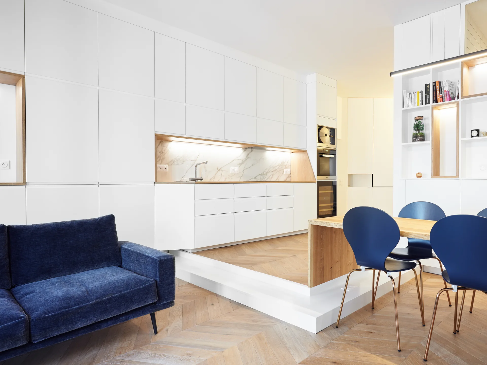 A modern open-concept space featuring a white minimalist kitchen on a raised wood platform, a blue sofa, and a matching blue dining set.