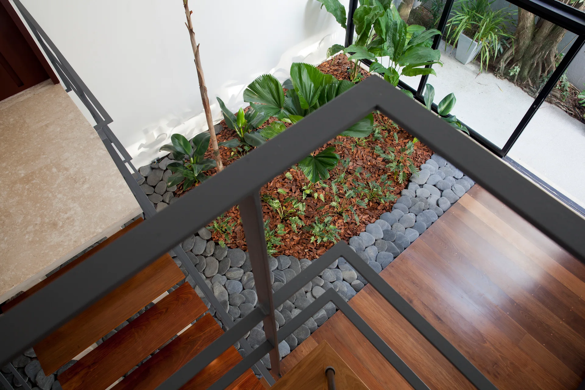 Looking down from a wooden staircase with a black metal railing onto an indoor garden bed filled with green plants, mulch, and a grey river stone border.