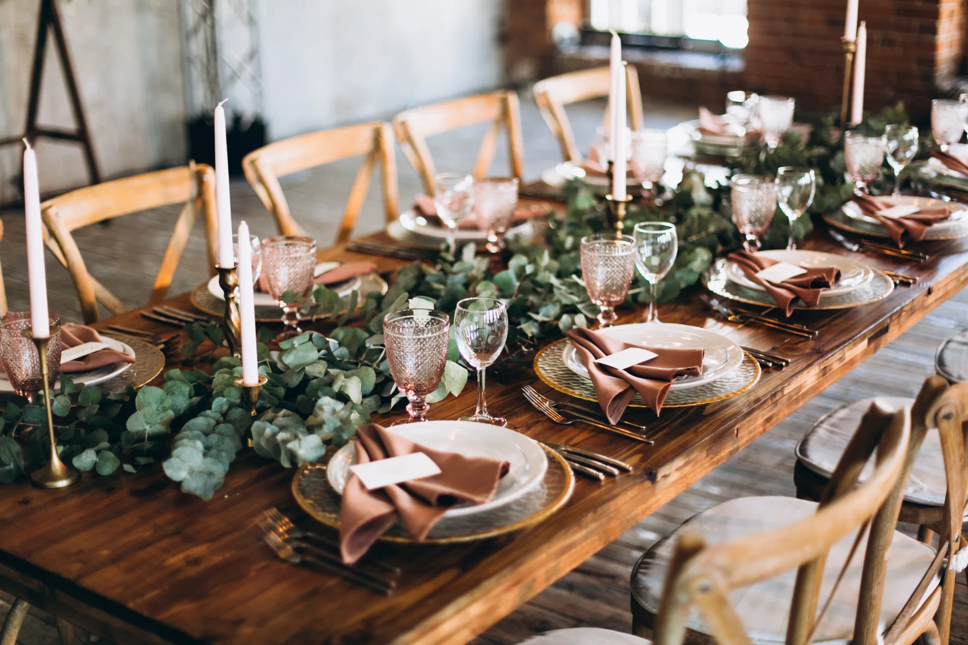 A long wooden dining table is set with a leafy green eucalyptus runner, tall taper candles, dusty rose napkins, and pink tinted glassware.