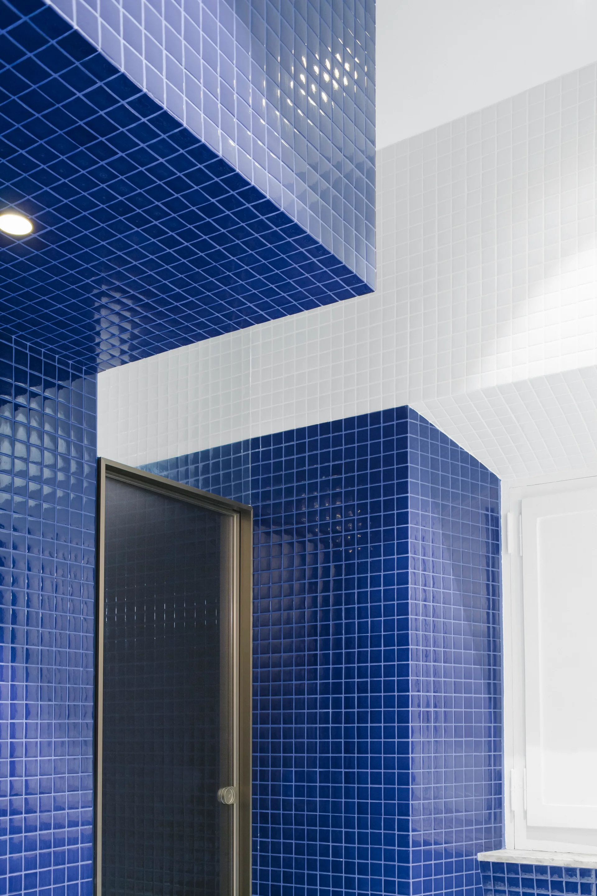 Glossy cobalt blue and bright white square tiles cover the walls and overhead soffit of a modern bathroom with a dark glass door.