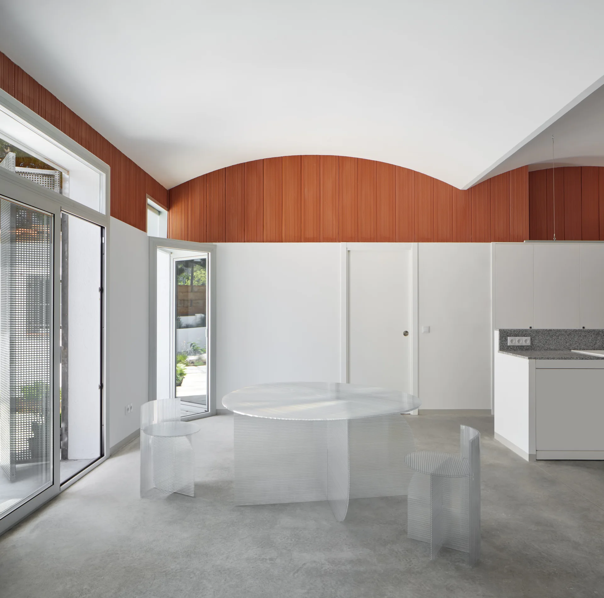 A minimalist dining space features a round transparent acrylic table and chairs on a concrete floor beneath a vaulted ceiling with terracotta panels.