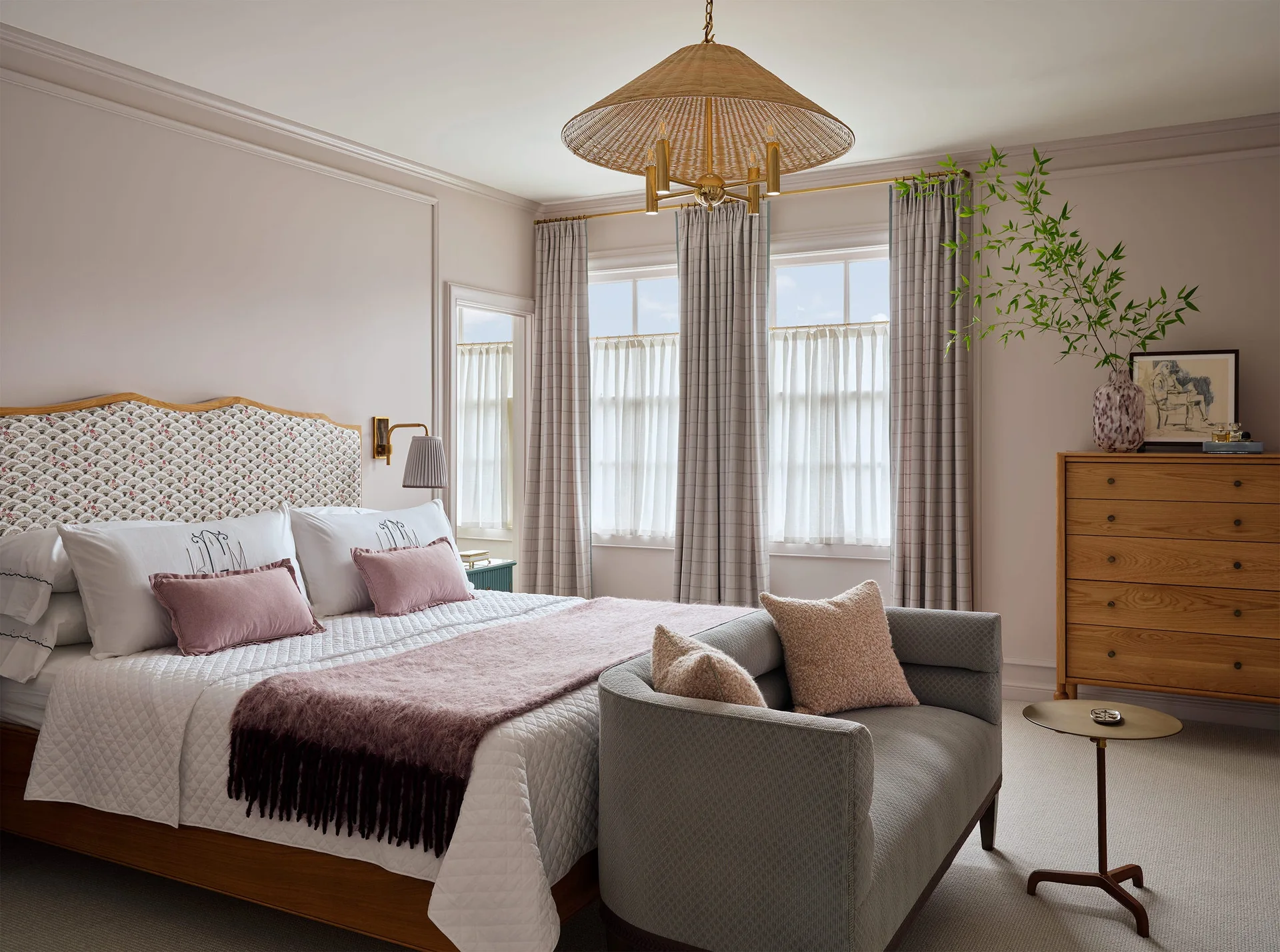 A bedroom features a bed with a patterned upholstered headboard, a grey footboard bench, and a woven rattan pendant light.