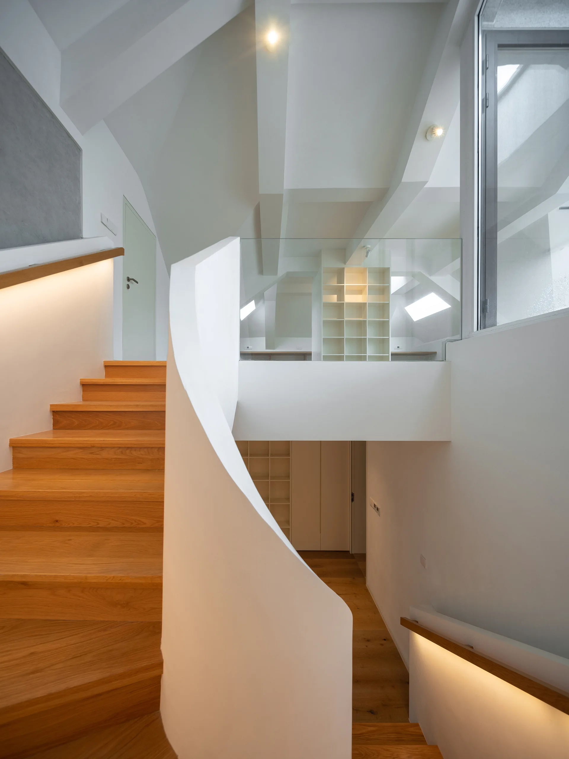 A modern staircase features light wood steps, a curved white solid railing, integrated handrail lighting, and a glass-walled landing.