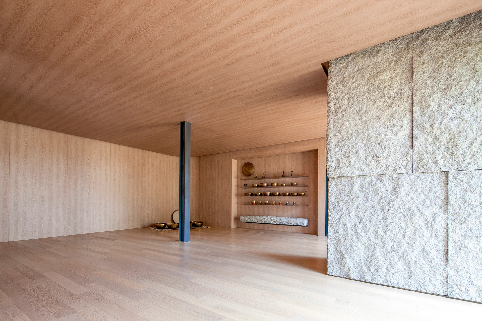 A minimalist wellness studio with light wood walls, floor, and ceiling, featuring a rough gray stone wall and recessed shelves displaying metal bowls.