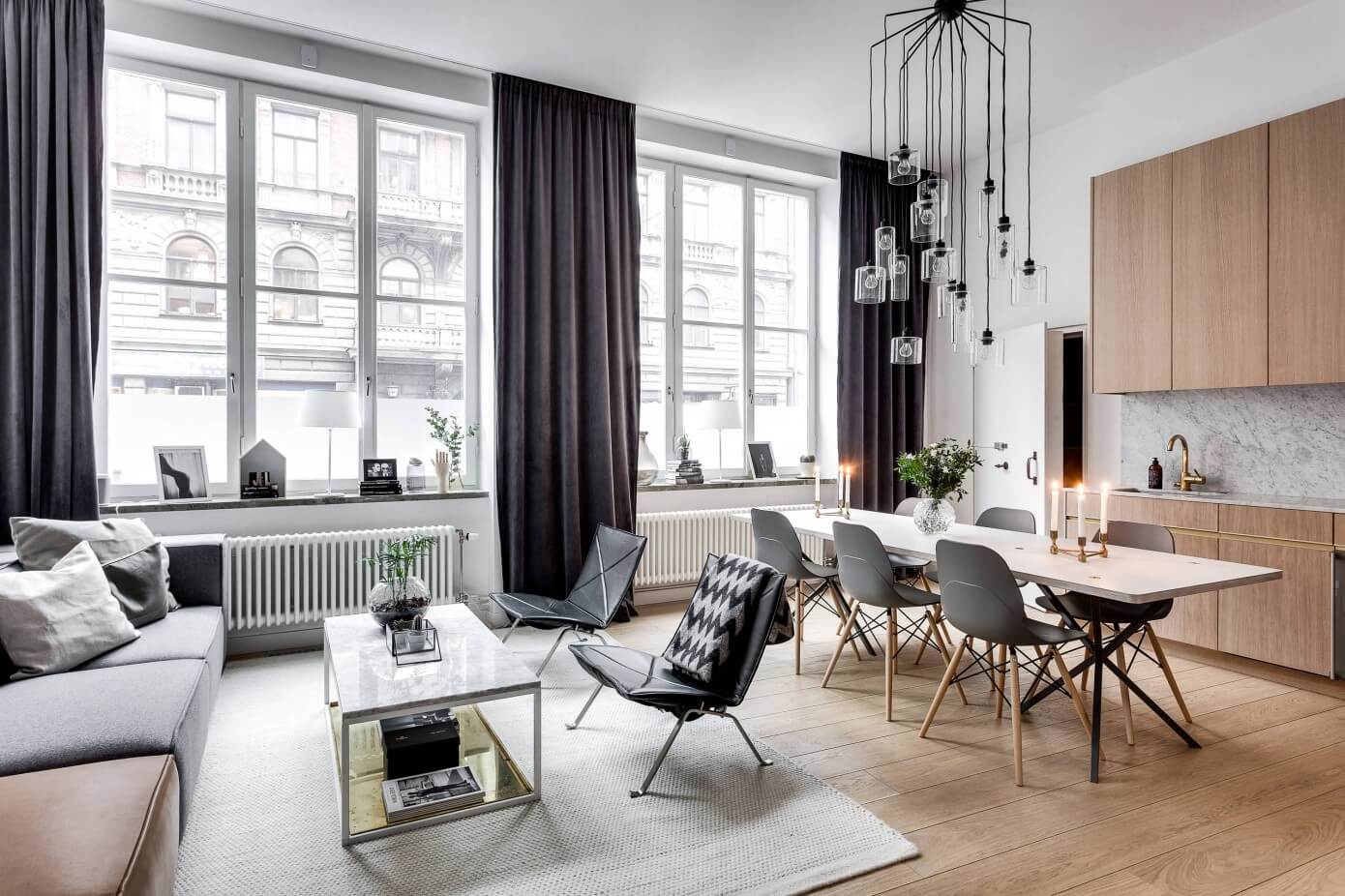 An open-plan living and dining room featuring a grey sofa, a long white table, a black multi-light pendant, and tall windows with dark drapes.