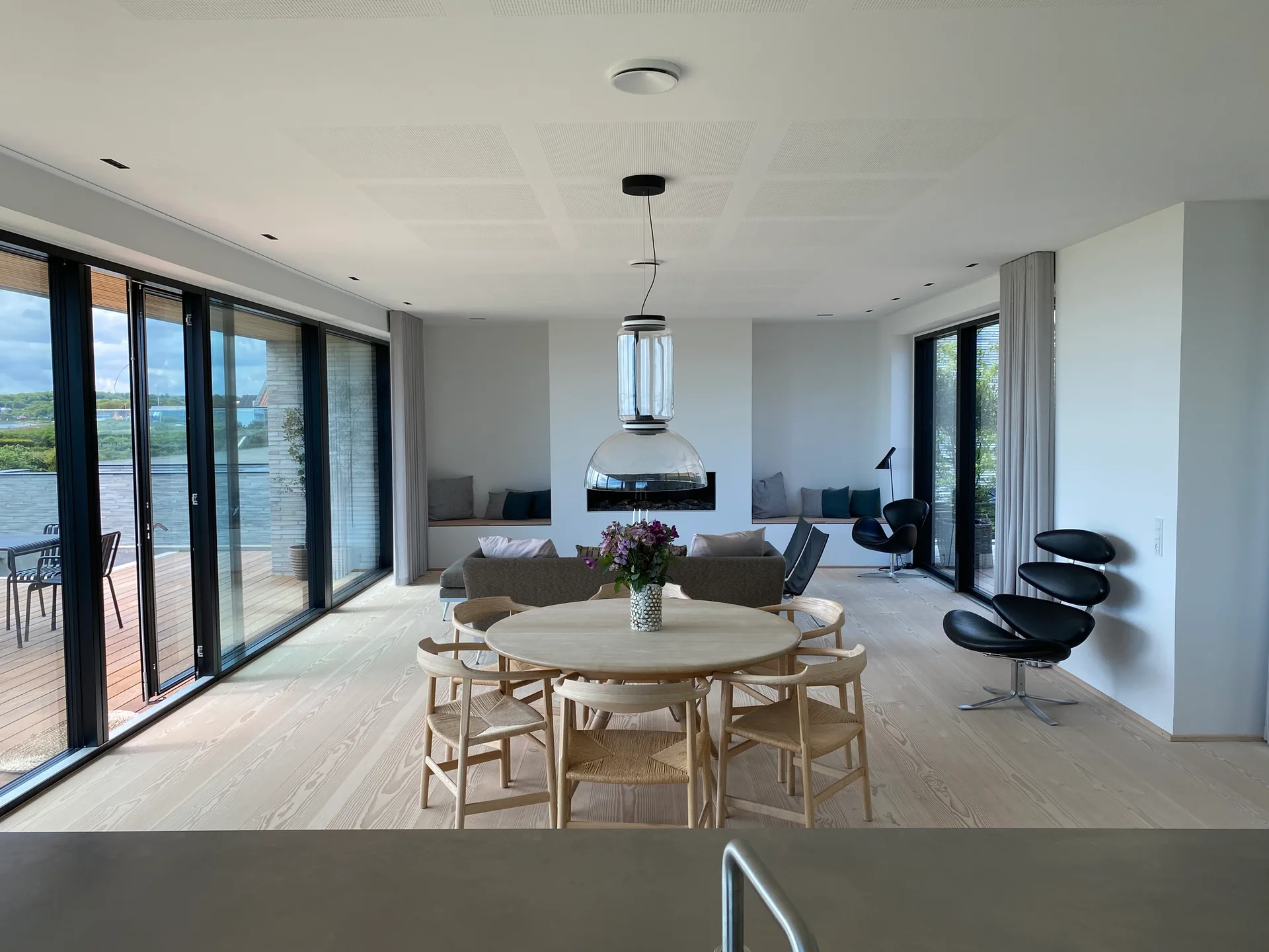 A light wood circular dining table and woven chairs sit under a clear glass pendant light in a bright, open-plan room with sliding glass doors.