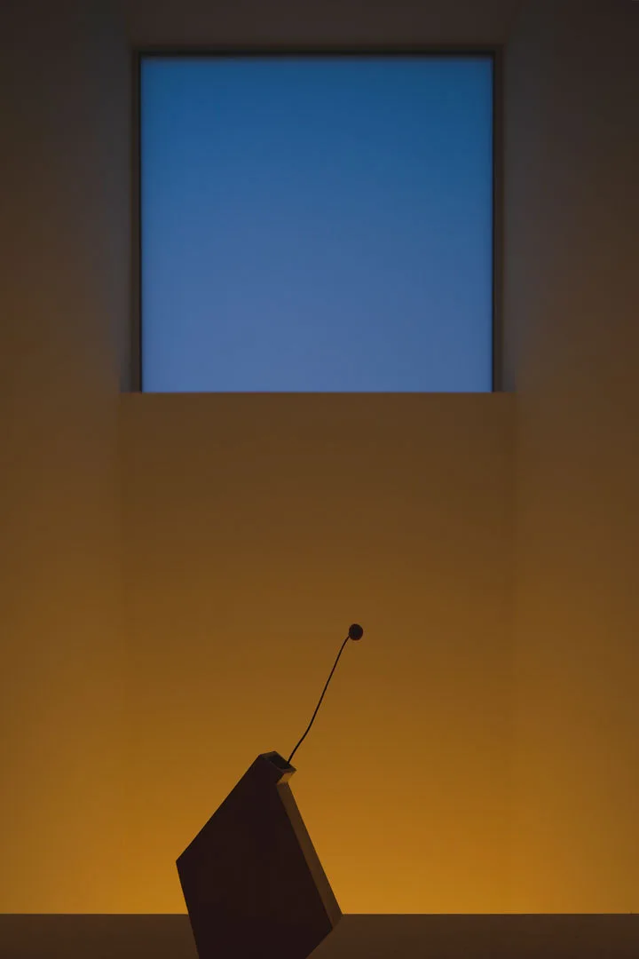 A dark angular sculpture sits in silhouette against a glowing yellow wall beneath a square skylight showing a bright blue sky.