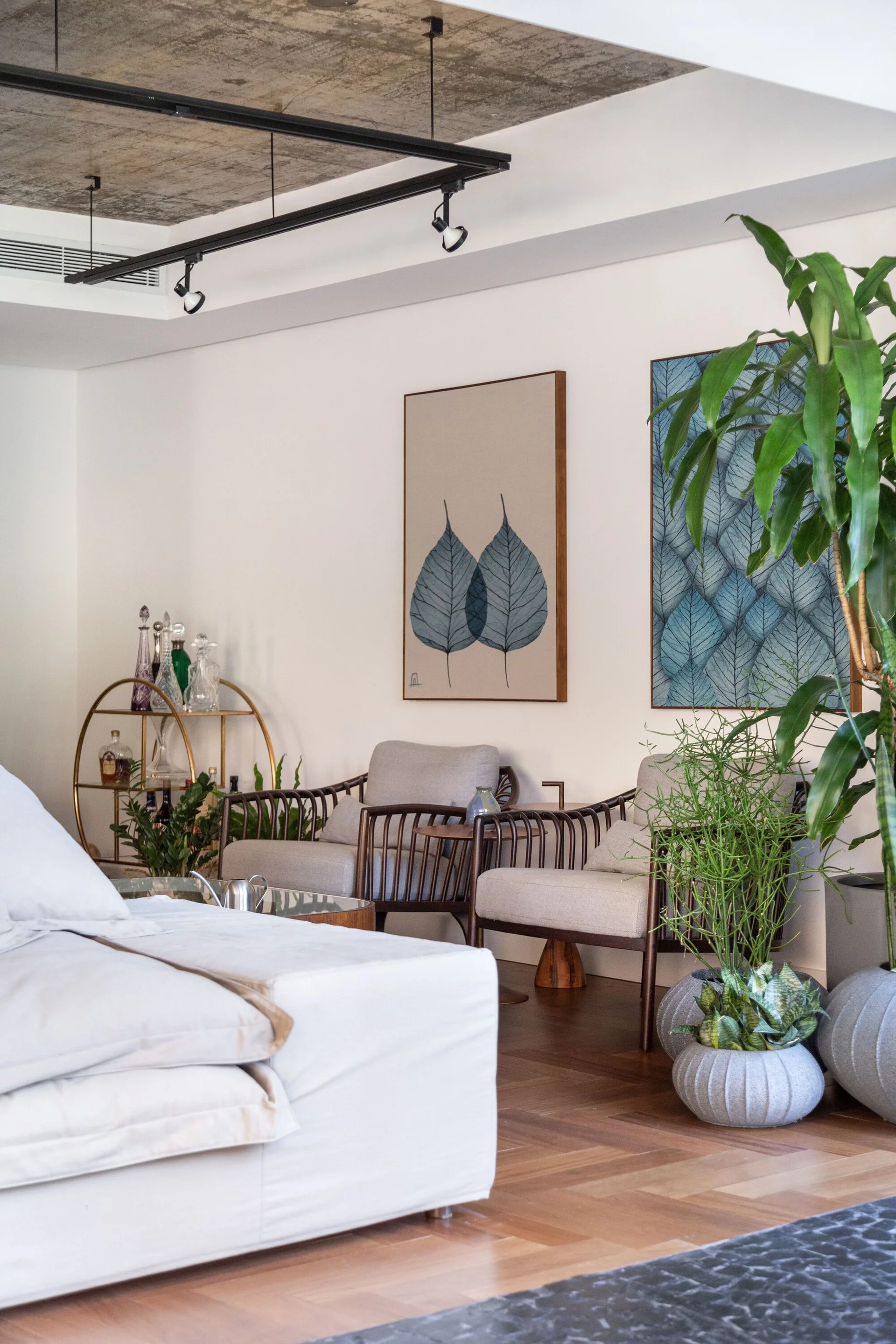 Two upholstered armchairs sit on parquet flooring below framed botanical art prints and an exposed concrete ceiling in a contemporary living room.