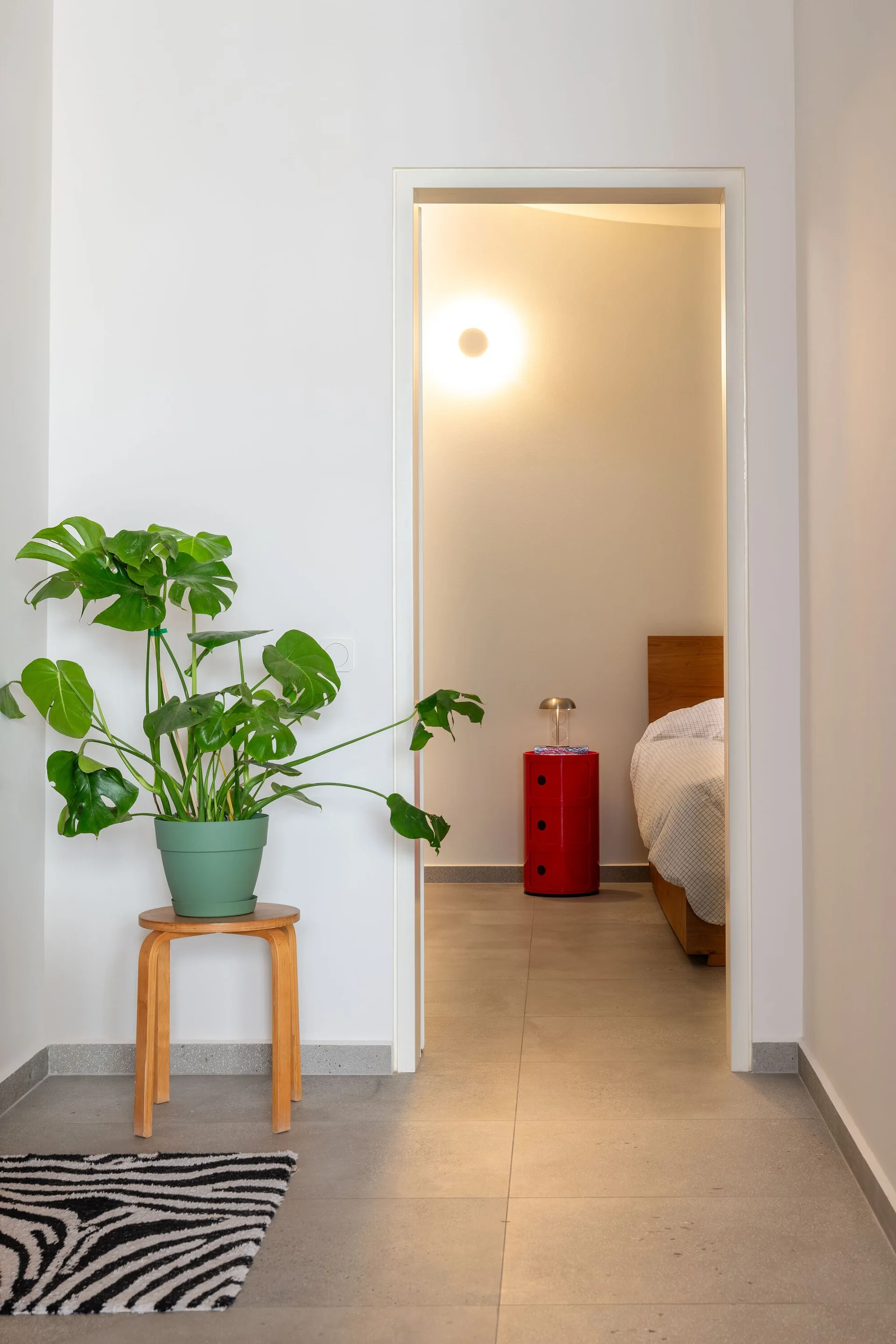 Looking through a doorway into a modern bedroom with a wooden bed, a red cylindrical nightstand, and a potted plant on a stool in the foreground.