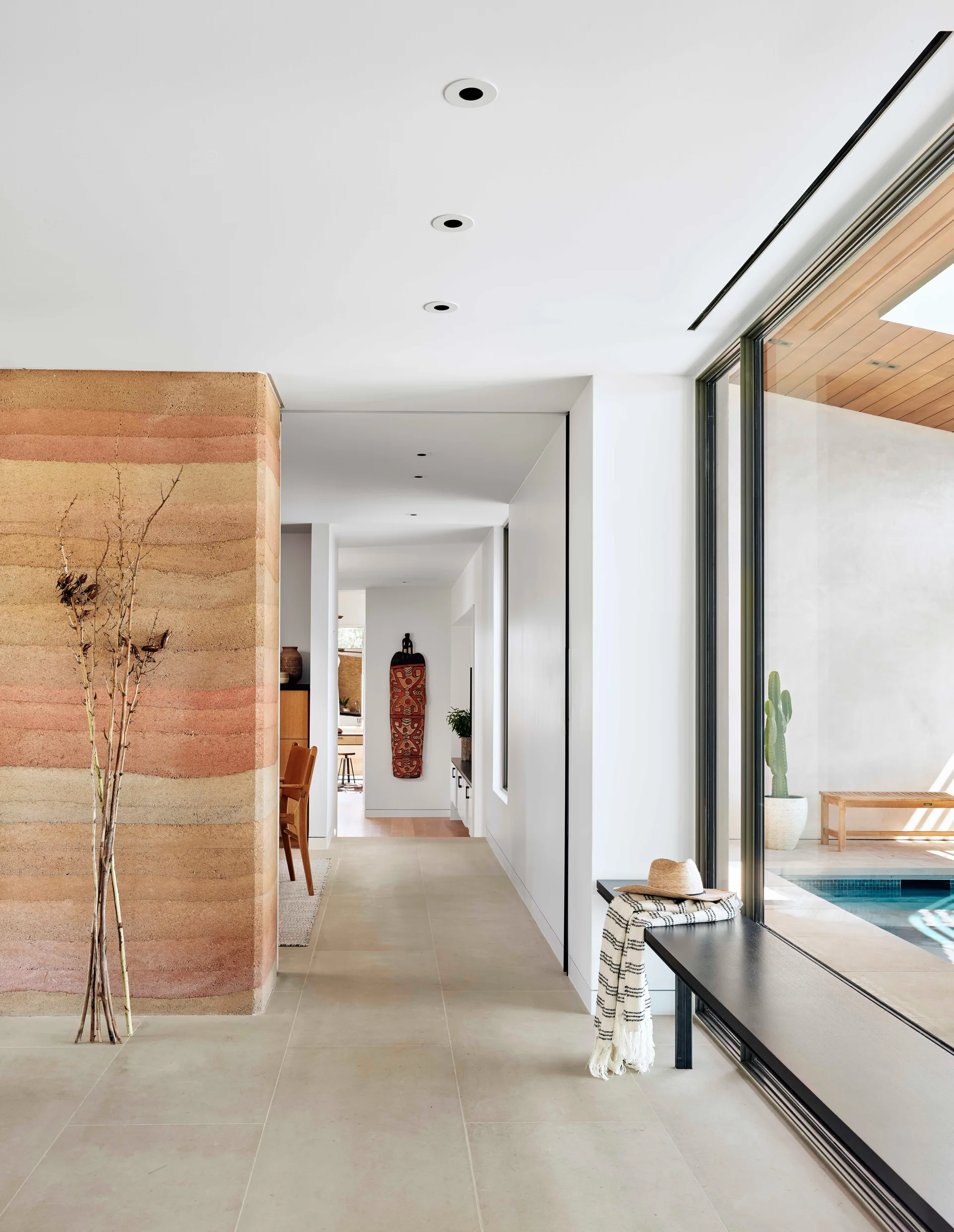 A bright hallway features a layered rammed earth wall in warm terracotta tones and a black bench next to a glass wall showing a pool.