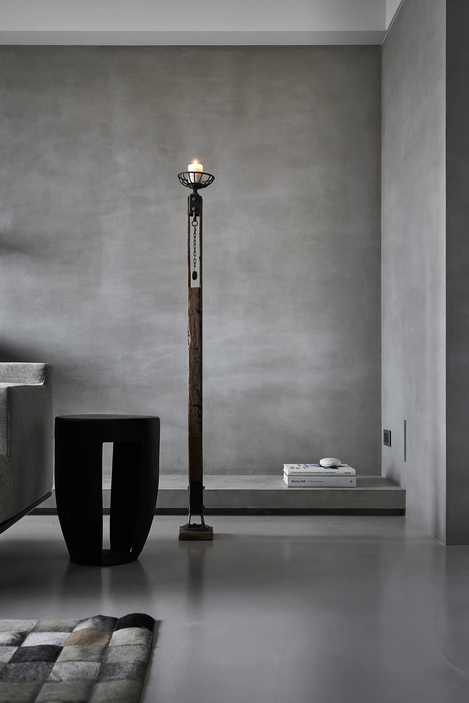 A minimalist living room corner featuring a tall weathered wood candle floor lamp and a dark sculptural side table against smooth concrete walls.