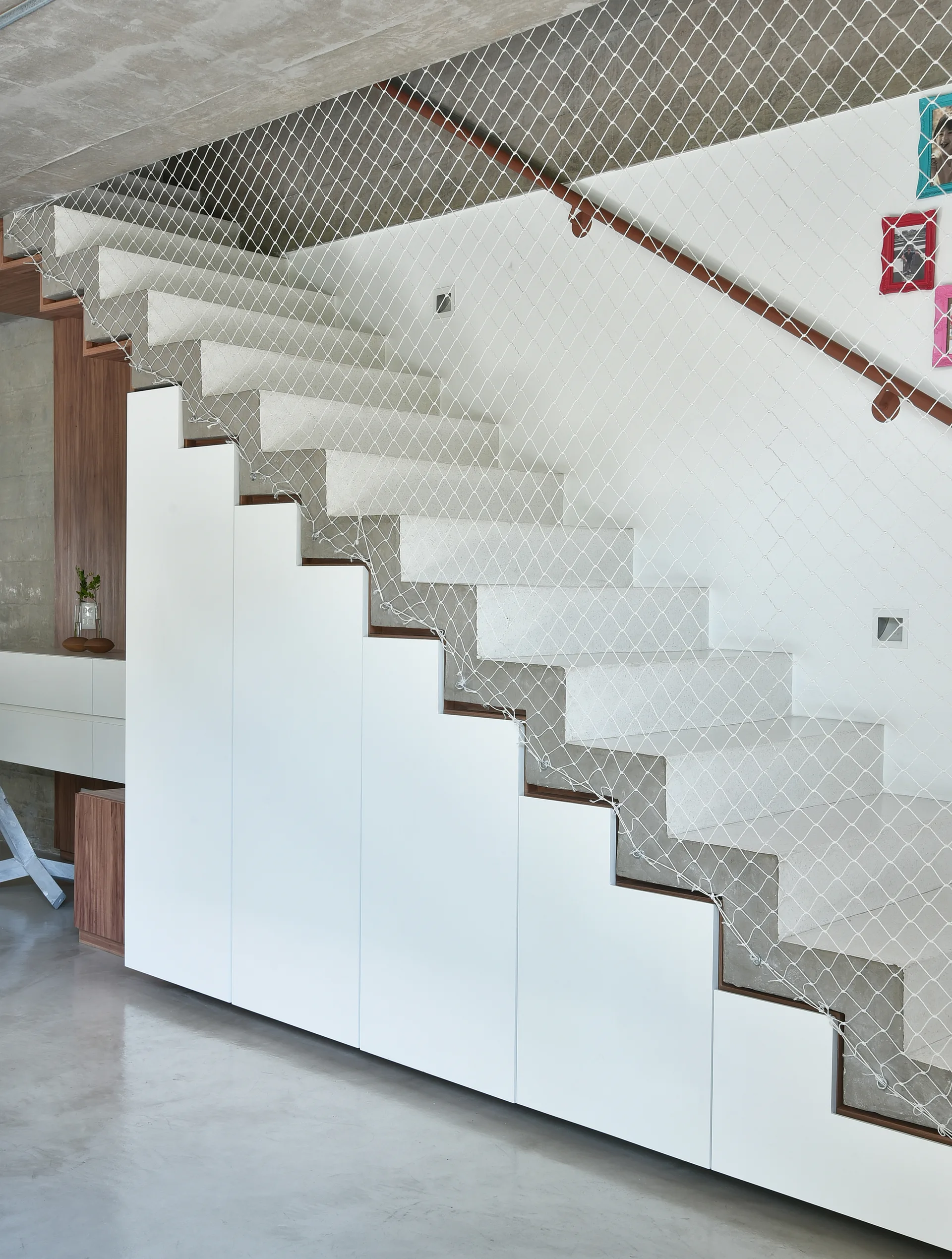 A modern white staircase features integrated under-stairs storage cabinets, a wooden handrail, and white mesh safety netting.