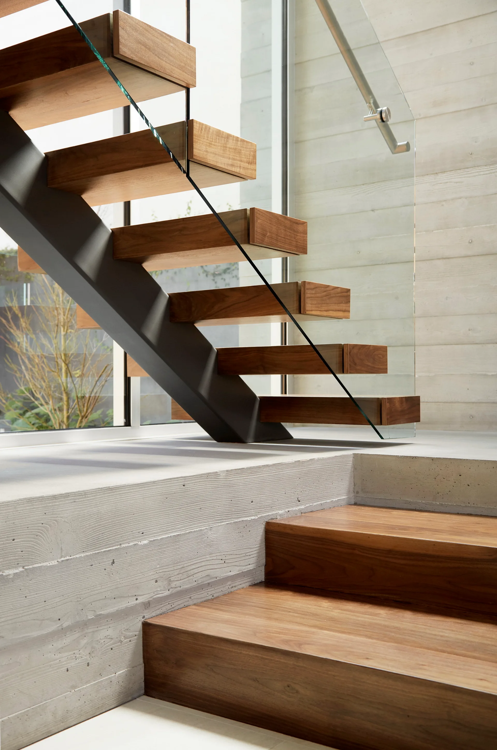 A minimalist floating staircase features thick wooden treads, a black metal support stringer, a glass railing, and a textured board-formed concrete wall.