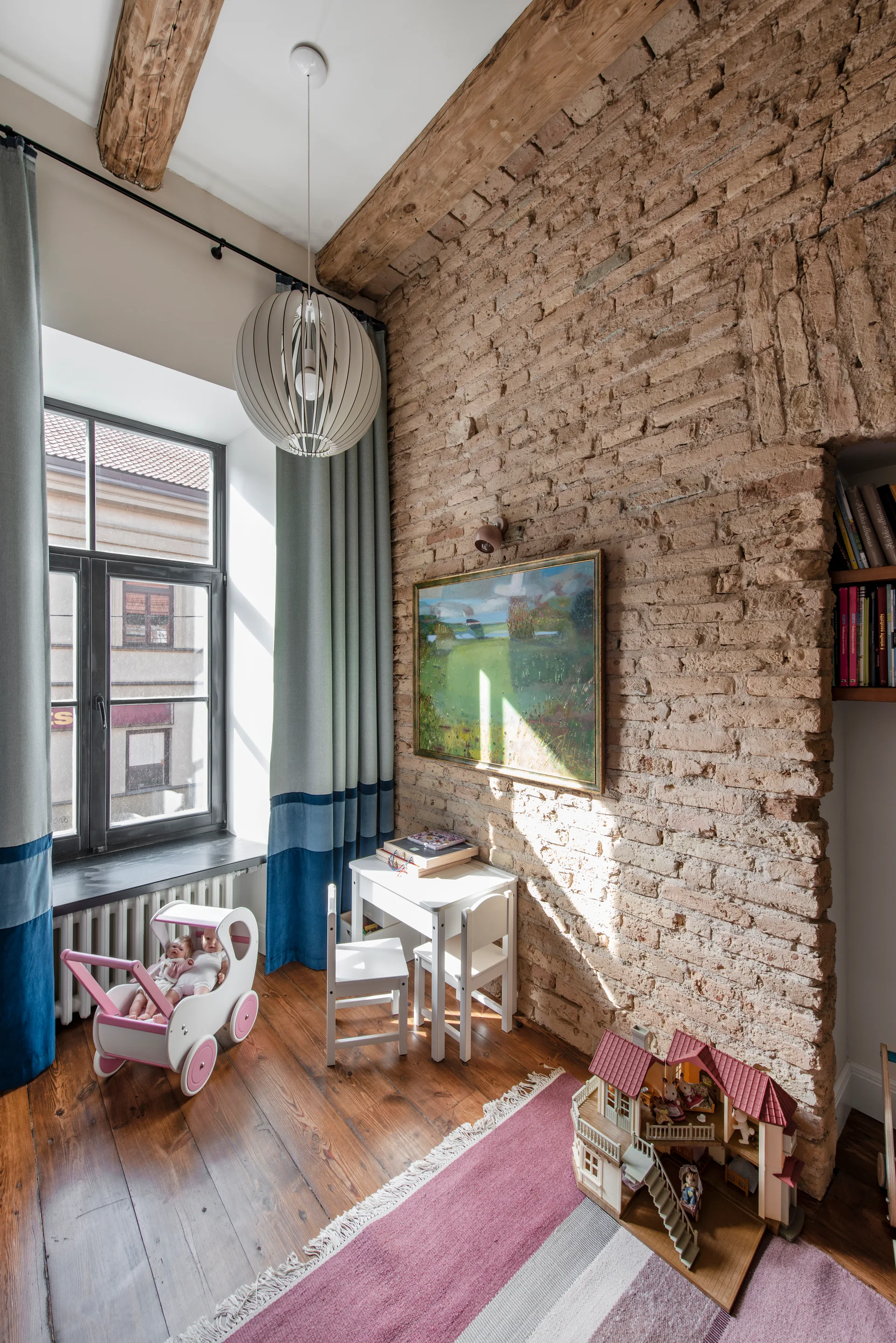 A bright children's playroom features an exposed brick wall, child-sized white furniture, color-blocked blue curtains, and wood flooring.
