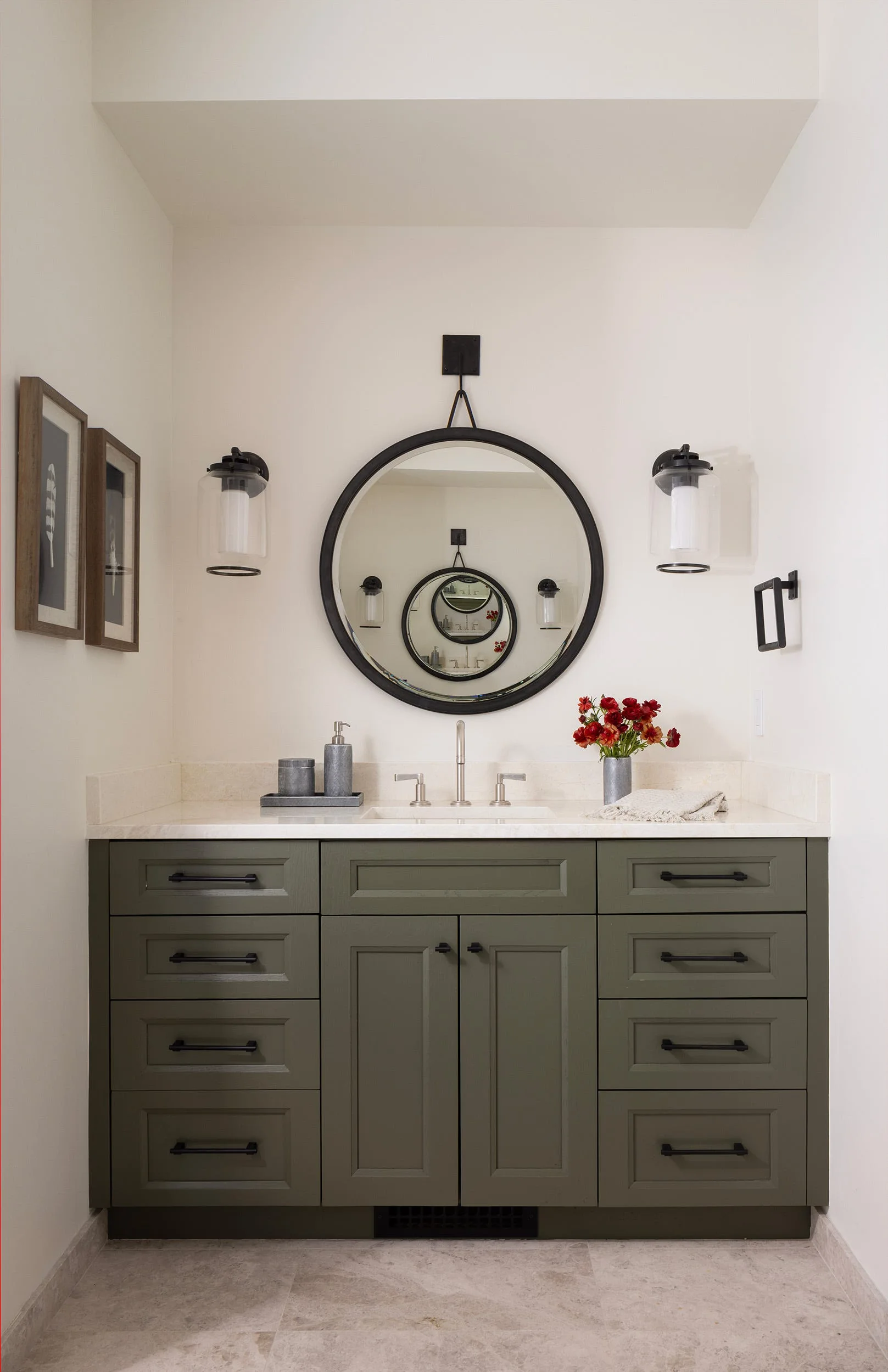 A sage green bathroom vanity with a light stone countertop, an undermount sink, and a round black-framed mirror flanked by glass sconces.