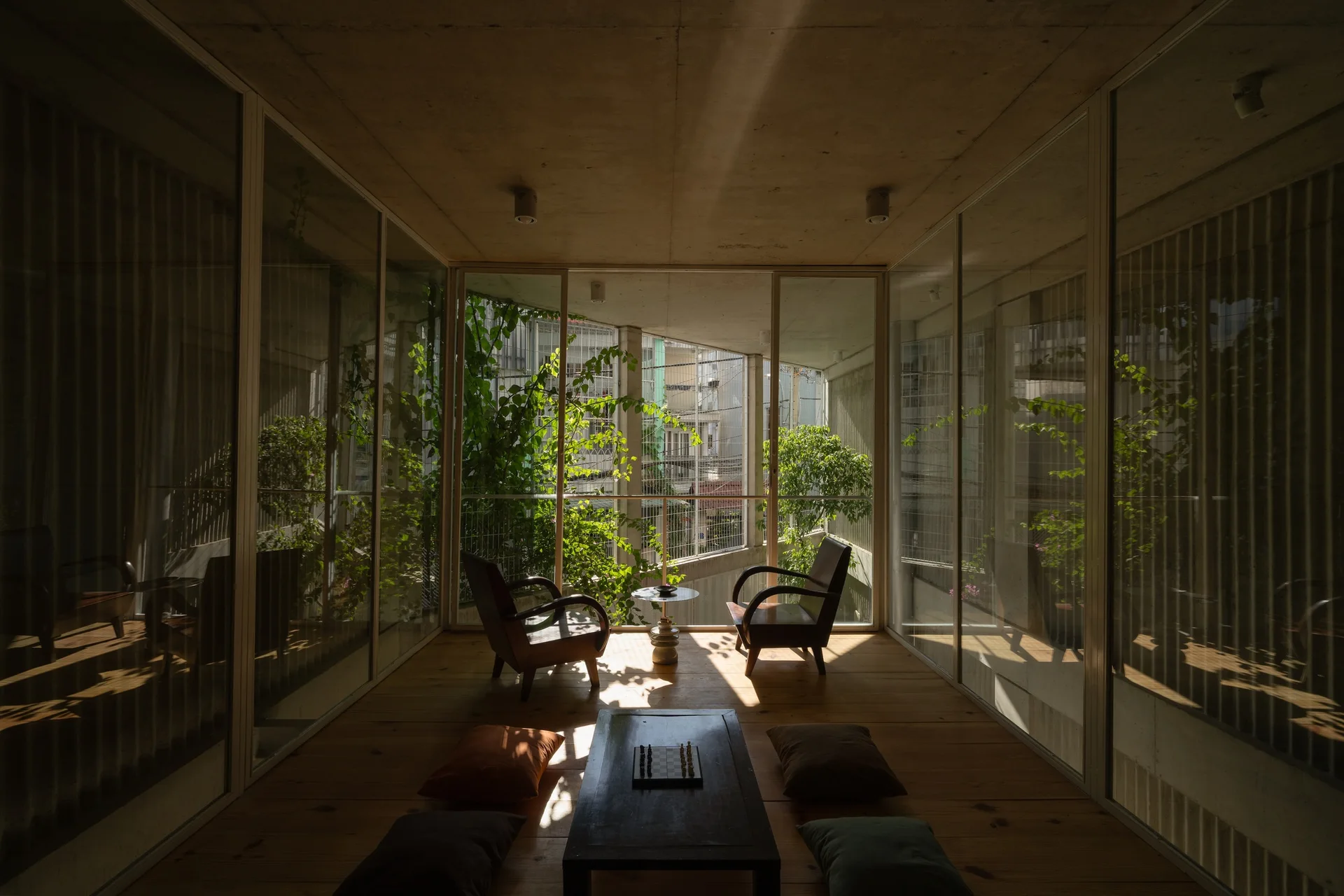 A minimalist sunroom features wood flooring, floor-to-ceiling glass walls, two wood armchairs, and a low coffee table with floor cushions.