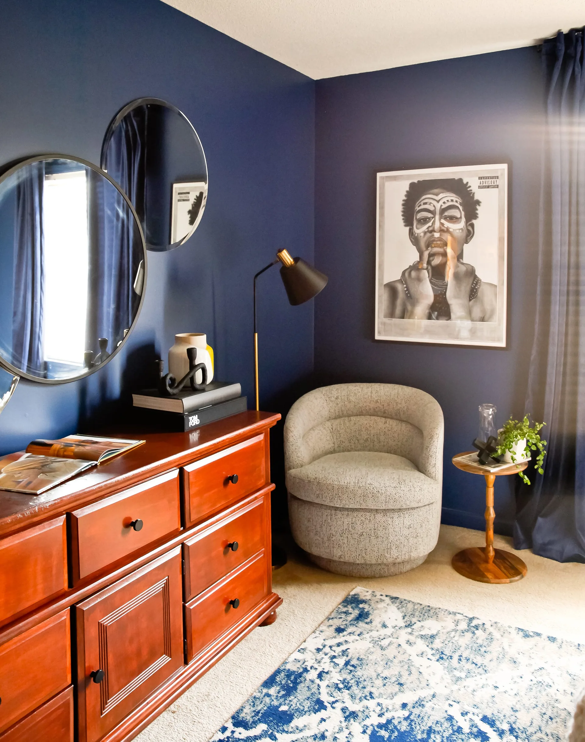 A bedroom corner features navy blue walls, a warm wood dresser with round mirrors, and a textured grey barrel armchair next to a small side table.
