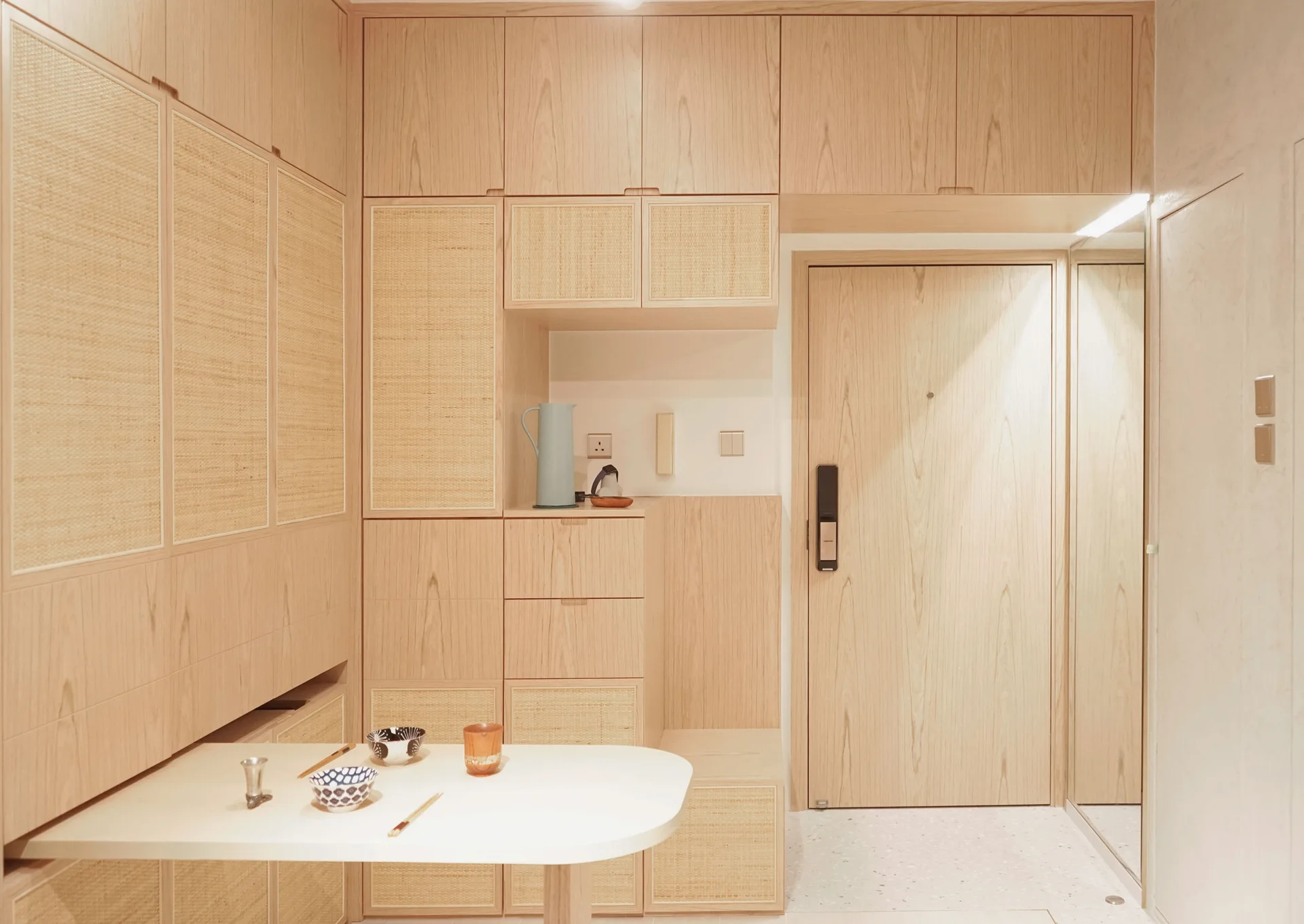 Floor-to-ceiling light wood cabinets with woven rattan inserts flank a wooden entryway door and an integrated white fold-out table.