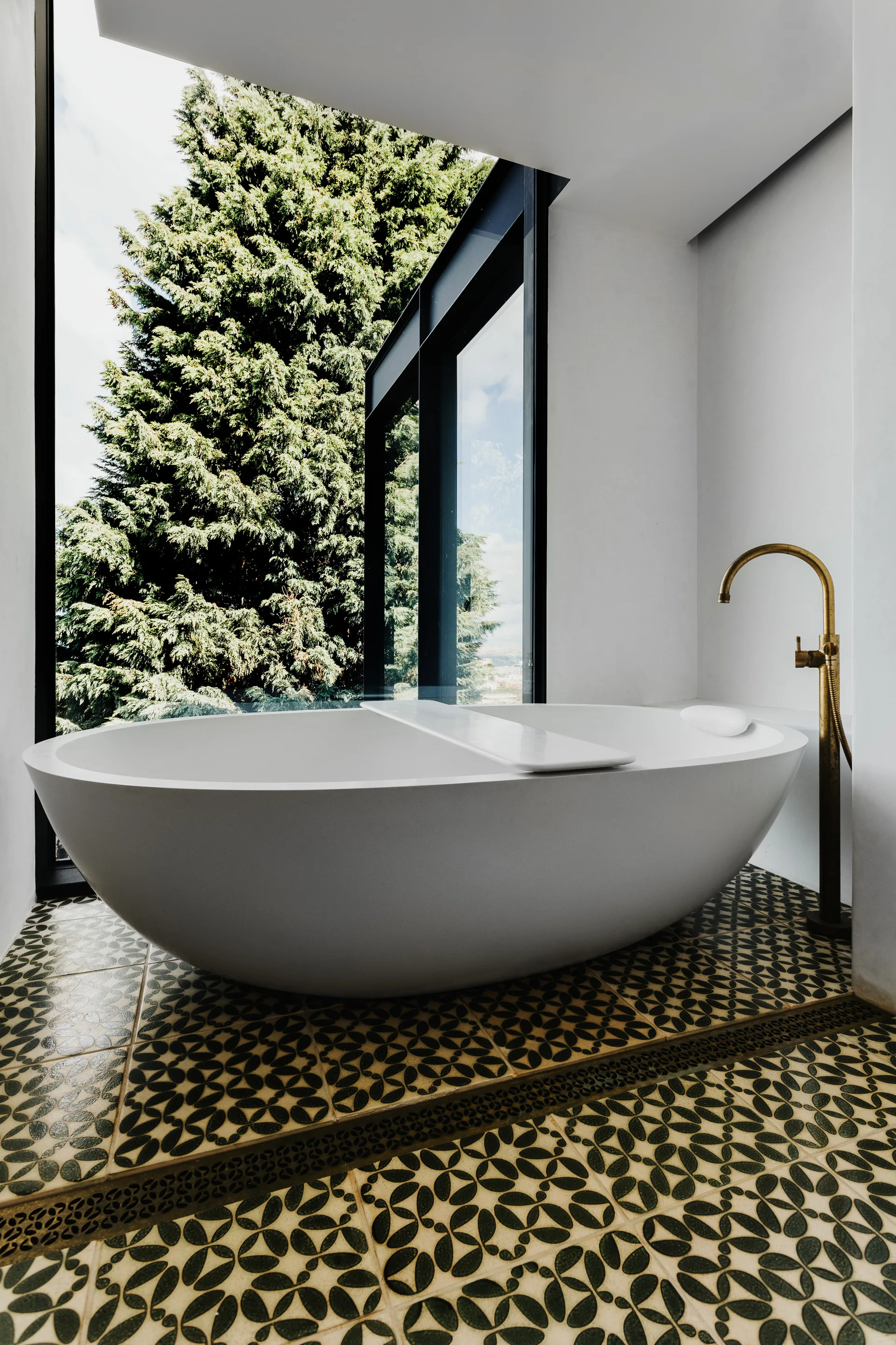 A white freestanding oval bathtub sits on green and white patterned tiles next to a tall glass window overlooking green trees and a brass floor faucet.