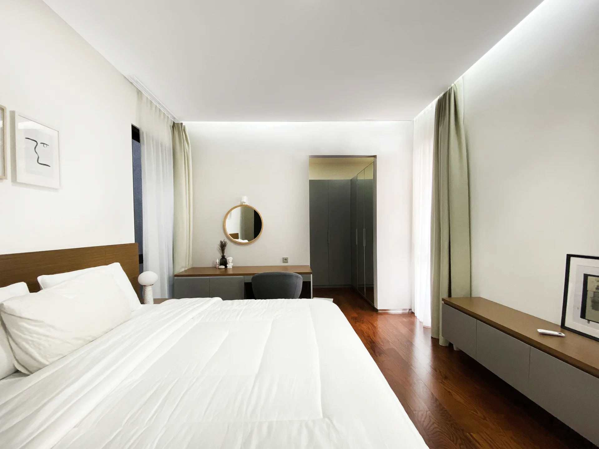 A minimalist bedroom featuring a white bed, dark wood floor, round mirror above a grey vanity, and light green floor-to-ceiling curtains.