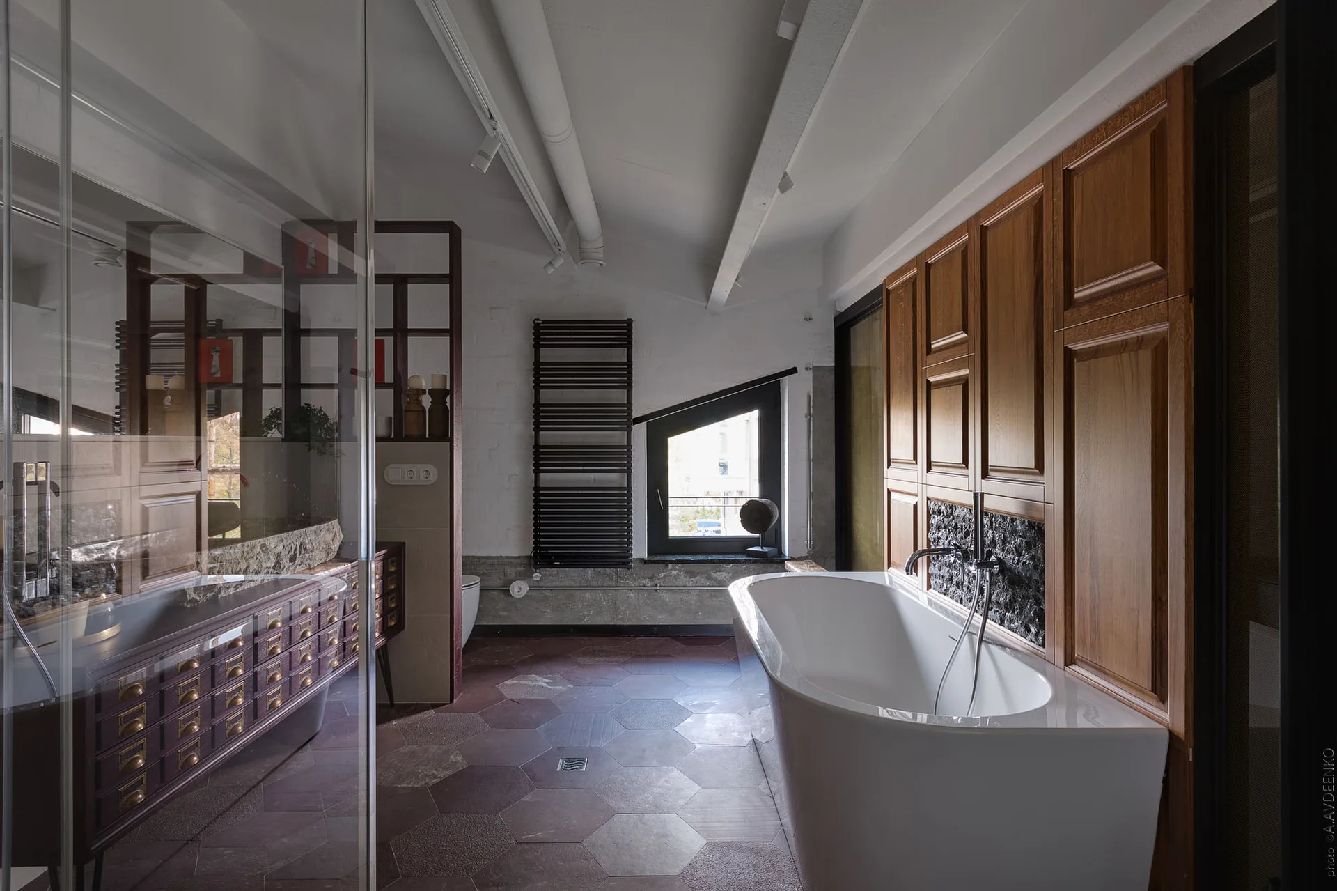 A modern bathroom features a white freestanding bathtub set against wood wall paneling, with terracotta hexagonal floor tiles and an angled window.