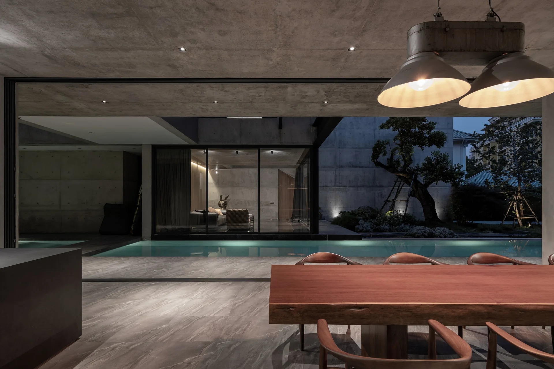 A modern dining room featuring a live-edge wooden table, grey stone floors, a raw concrete ceiling, and a view of a long indoor swimming pool.