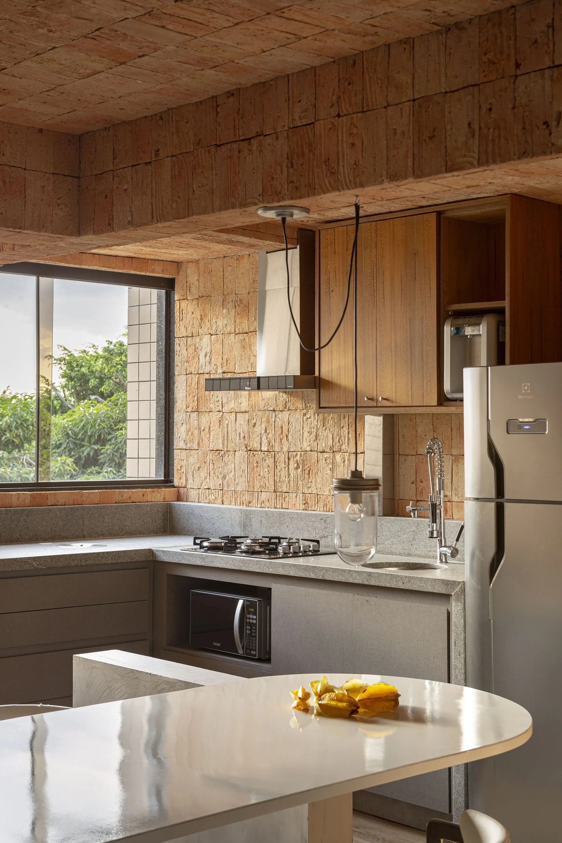 A modern kitchen features exposed terracotta brick walls and ceilings, grey stone countertops, wooden upper cabinetry, and a glossy white dining table.