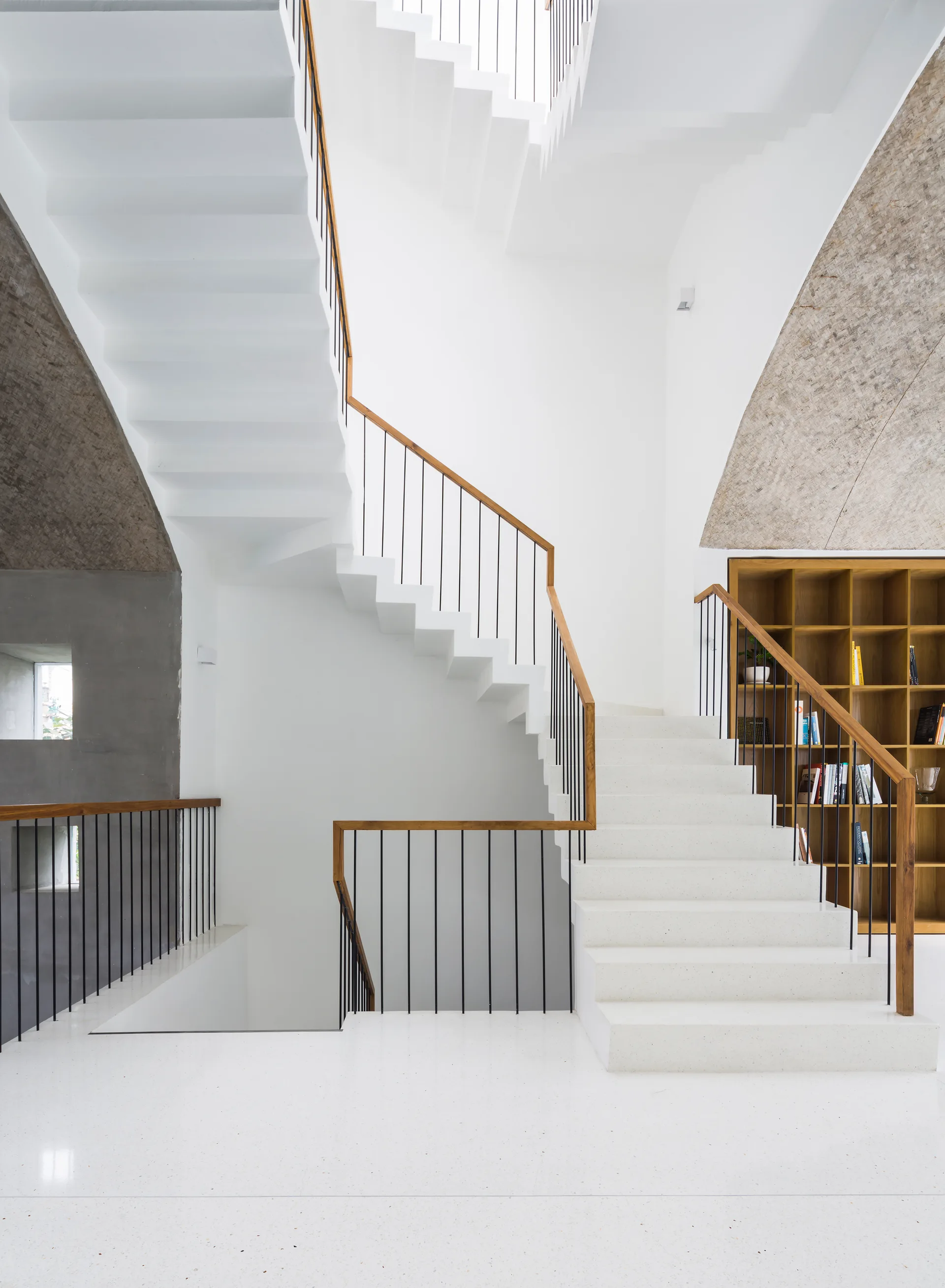A multi-level white staircase features thin black metal railings, a wooden handrail, and an integrated wood bookshelf next to a textured stone arch.