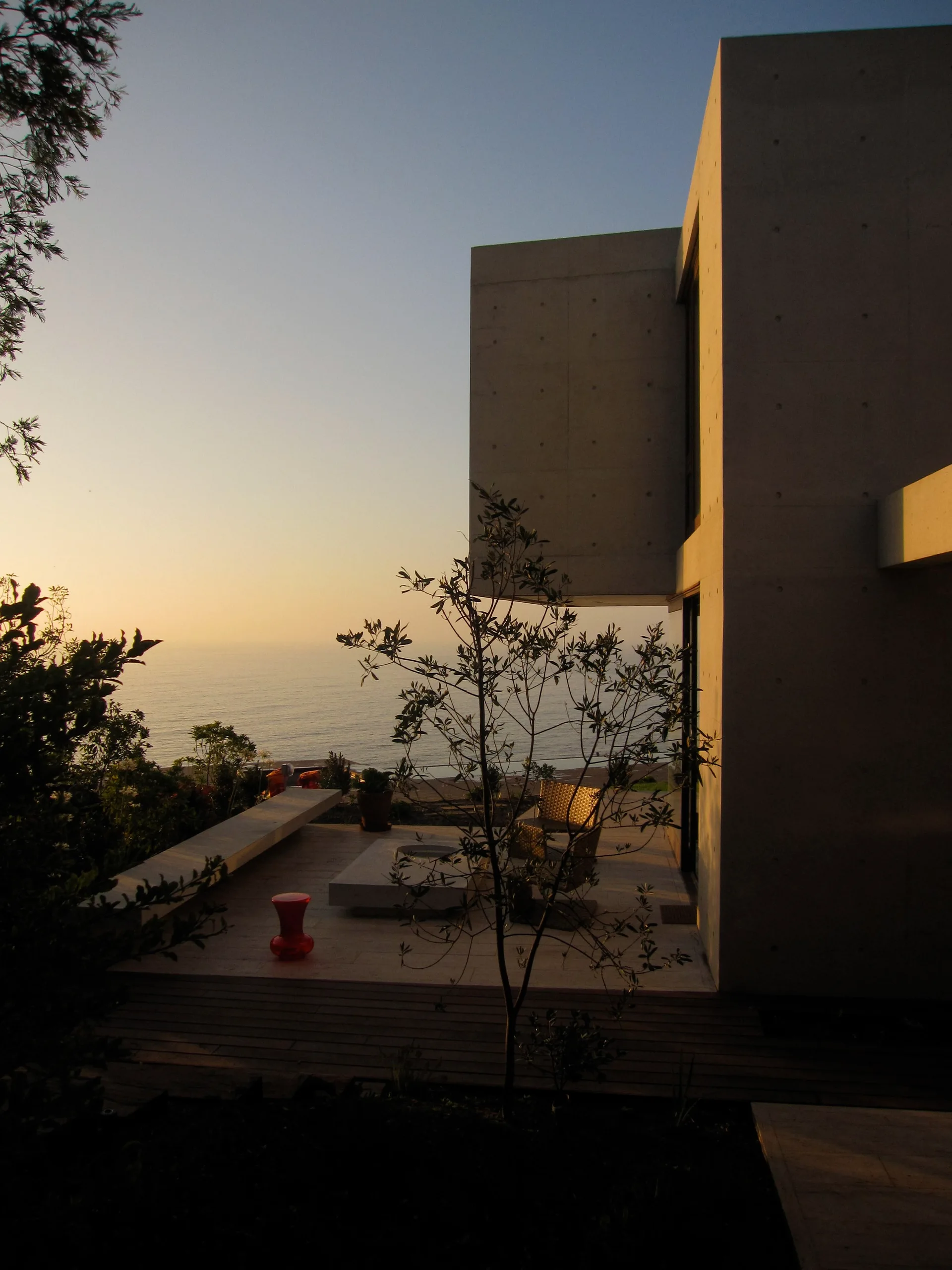 A modernist concrete building features a seaside patio with a wood deck, a small red stool, and a simple chair overlooking the ocean at sunset.