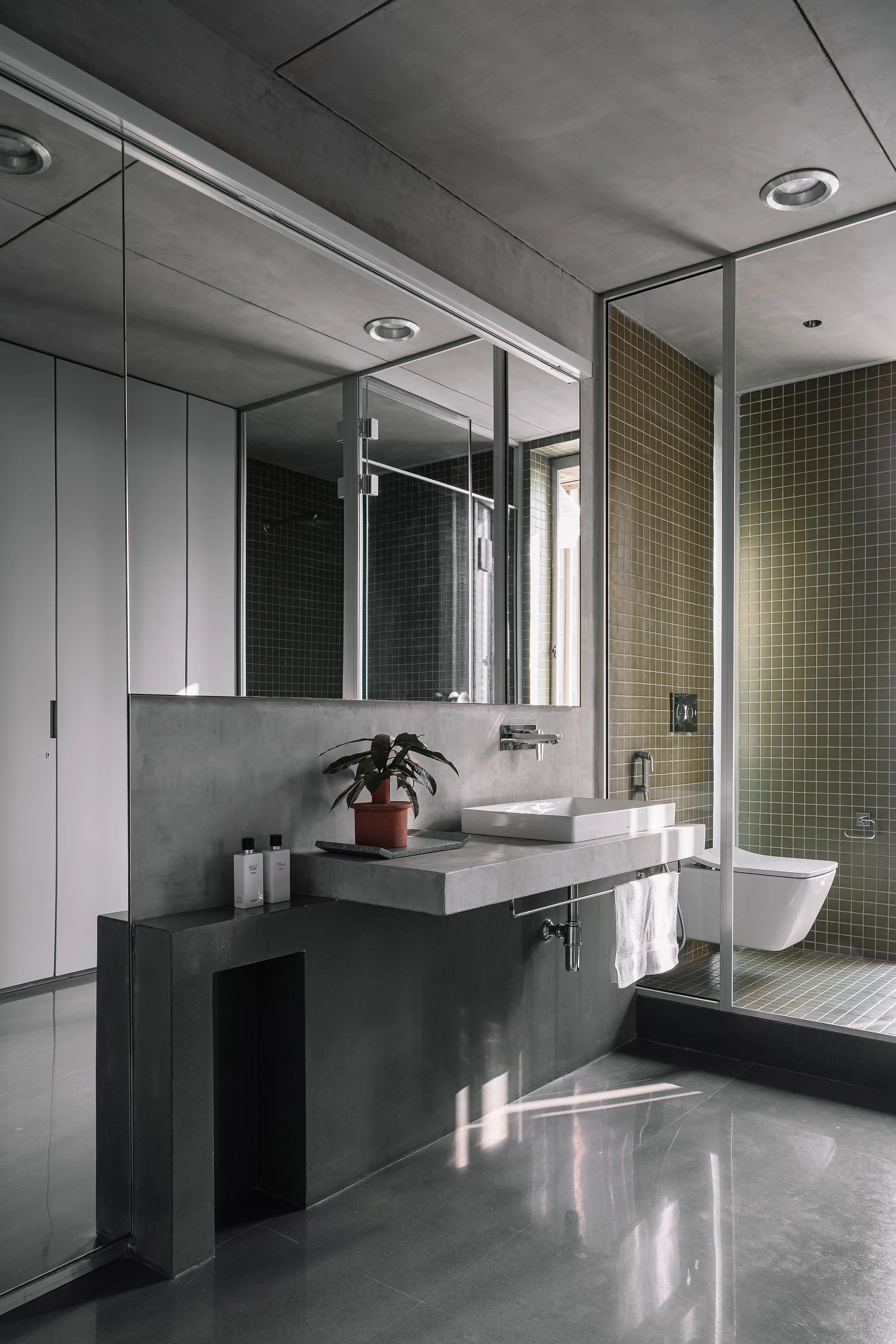 A modern bathroom features a floating concrete vanity with a white vessel sink beneath a large mirror, next to a shower lined with olive green square tiles.