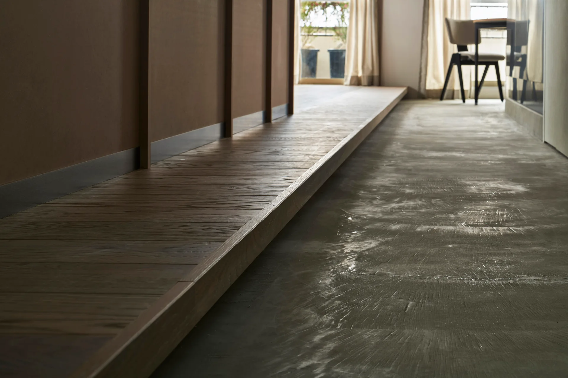 A close-up view of a raised brown wood platform transitioning to a textured grey concrete floor, with a dining area visible in the background.