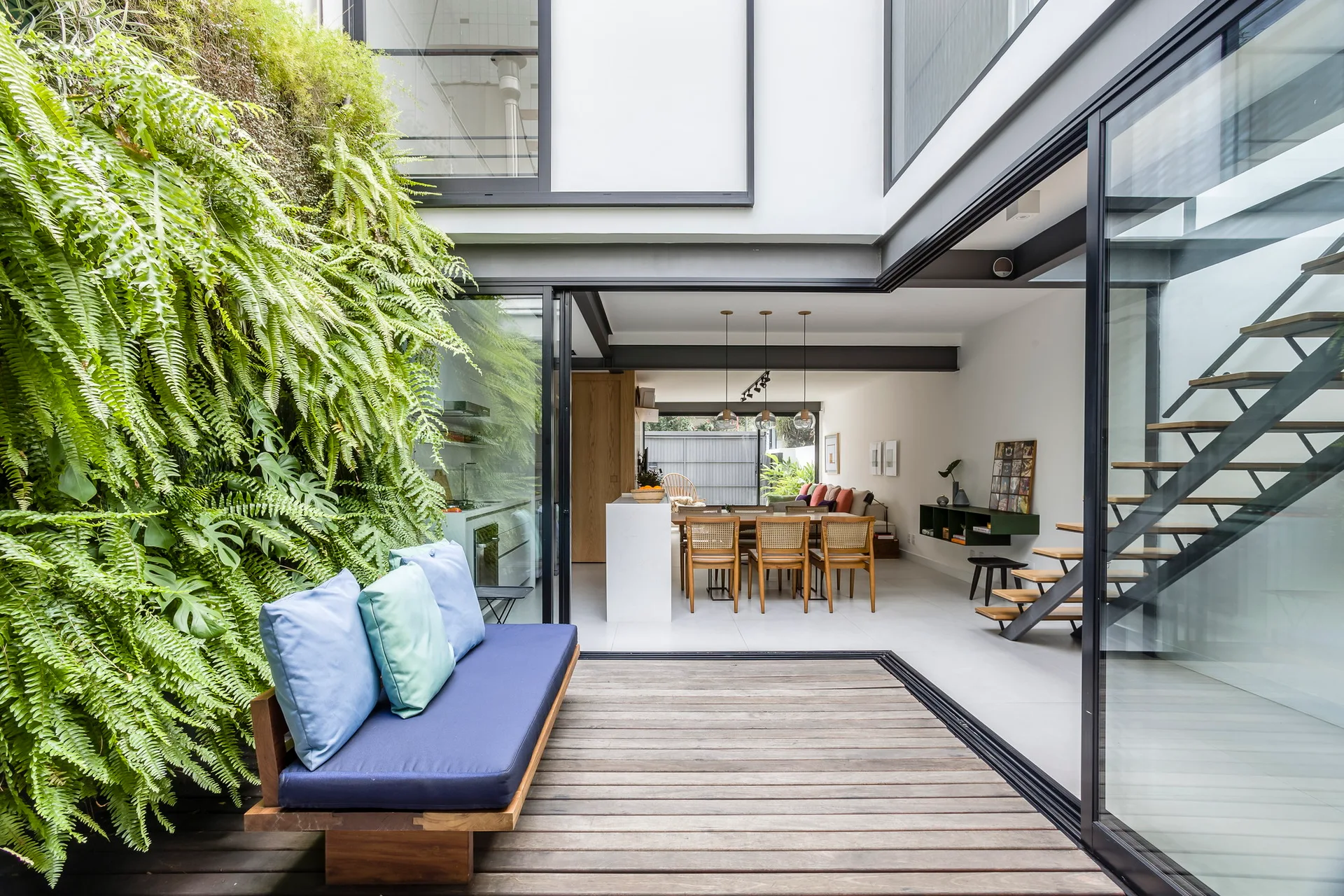 An open-concept living area connects to an outdoor wooden deck featuring a lush vertical garden, blue bench, and an open riser staircase.