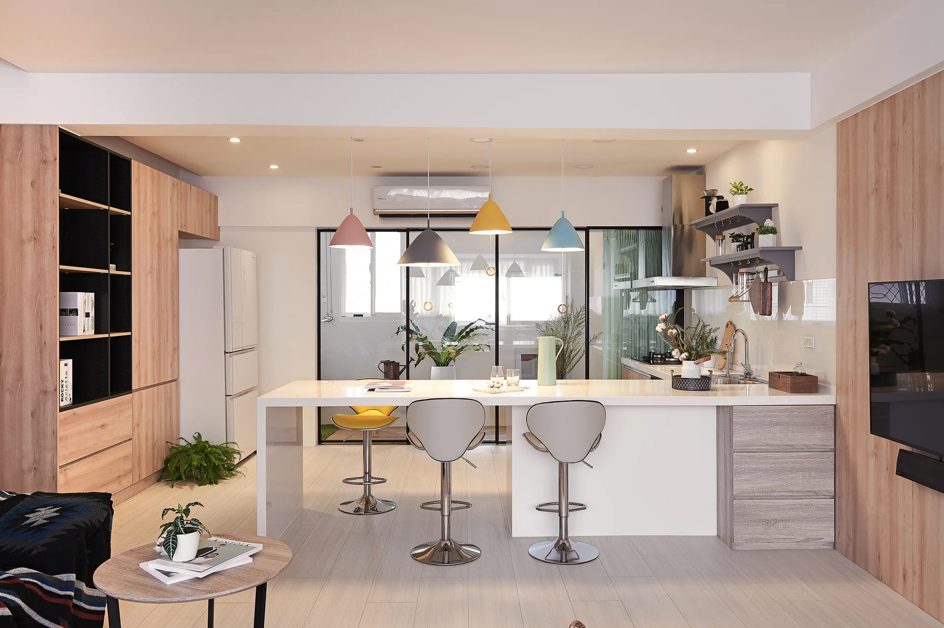 A modern kitchen features a white island with grey bar stools, four colorful pastel pendant lights, and light wood grain cabinetry.