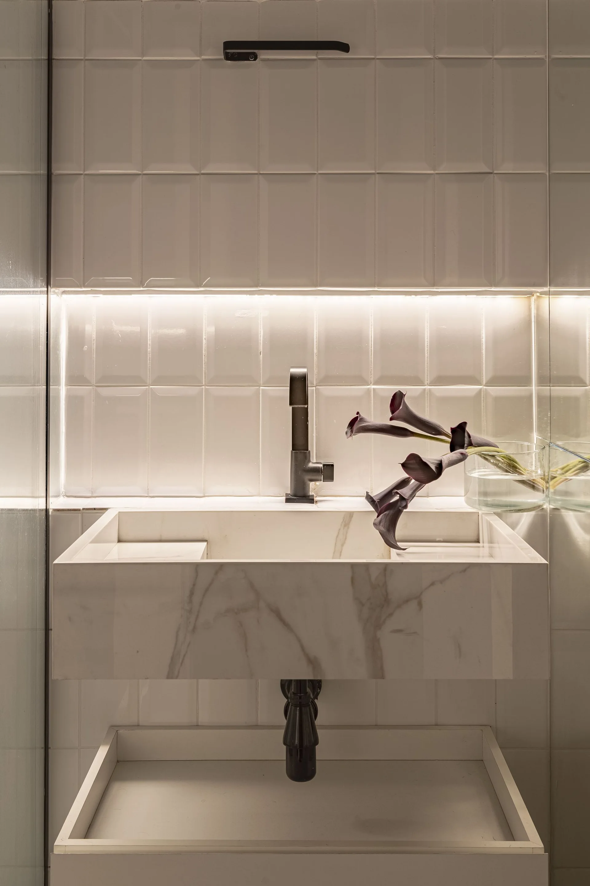 A wall-mounted white marble bathroom sink with a dark faucet, set against vertically stacked subway tiles and a glowing horizontal backlight.