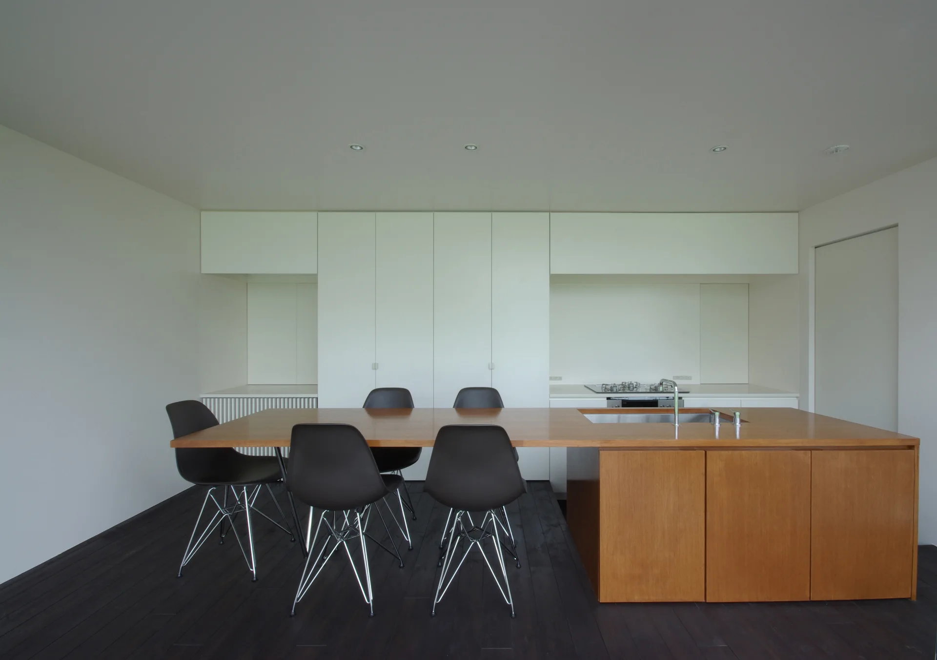 A minimalist kitchen featuring a wood island with a sink and integrated dining table, black chairs, white cabinets, and dark flooring.