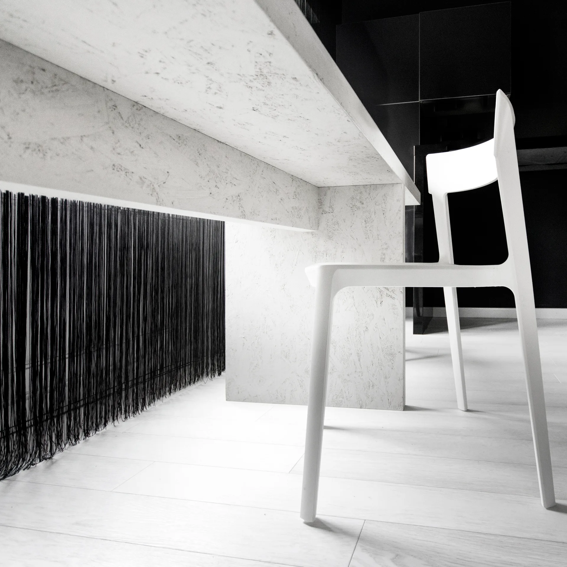 A close-up view of a mottled white table and a modern white chair set against a backdrop of long black fringe and a dark wall.