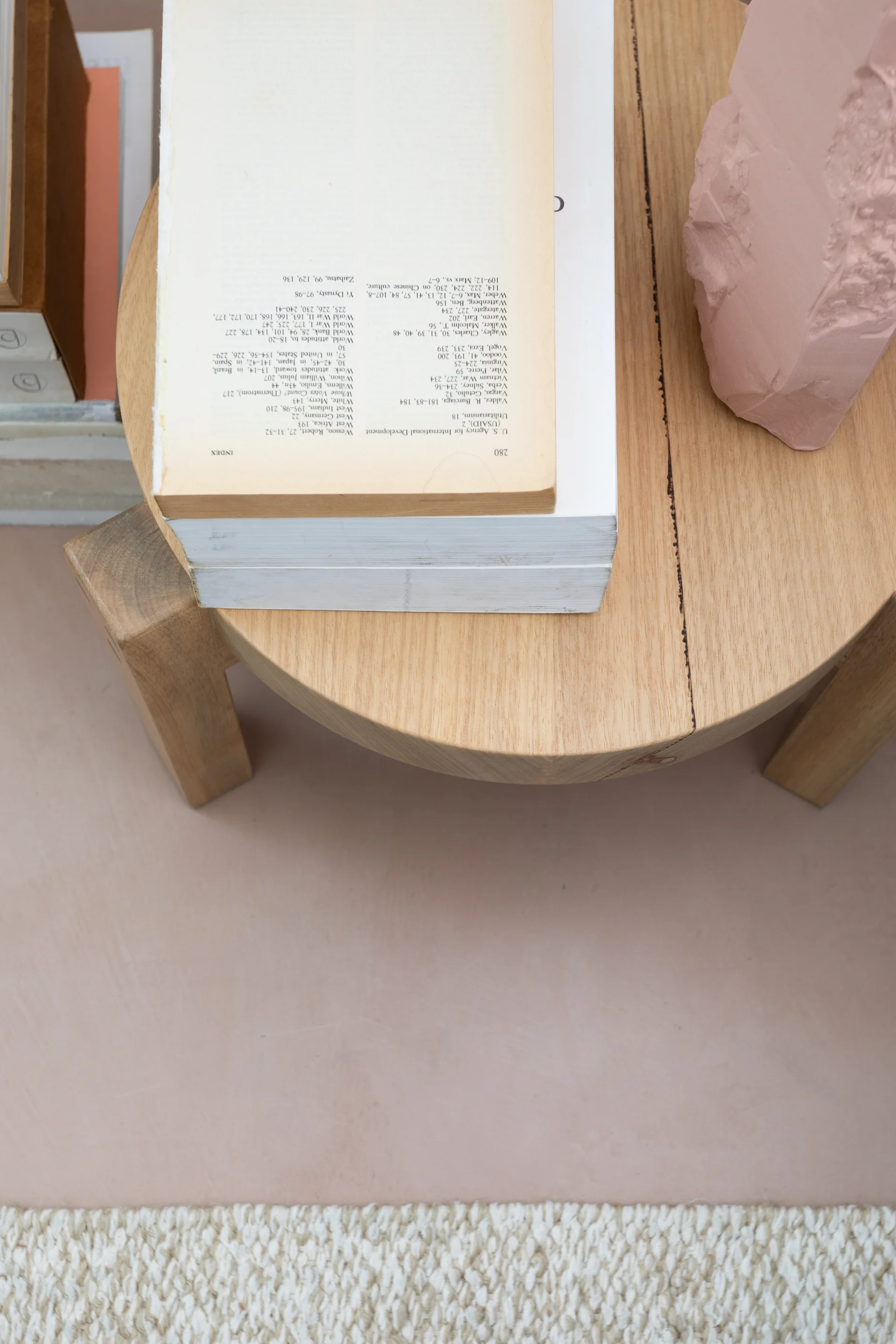 A top-down view of a light wood side table styled with stacked books and a textured pink sculpture, set over a neutral woven rug.