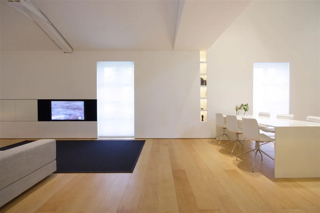 A minimalist open living room features a white dining set, light wood floors, integrated wall shelving with built-in lighting, and a flat-screen television.