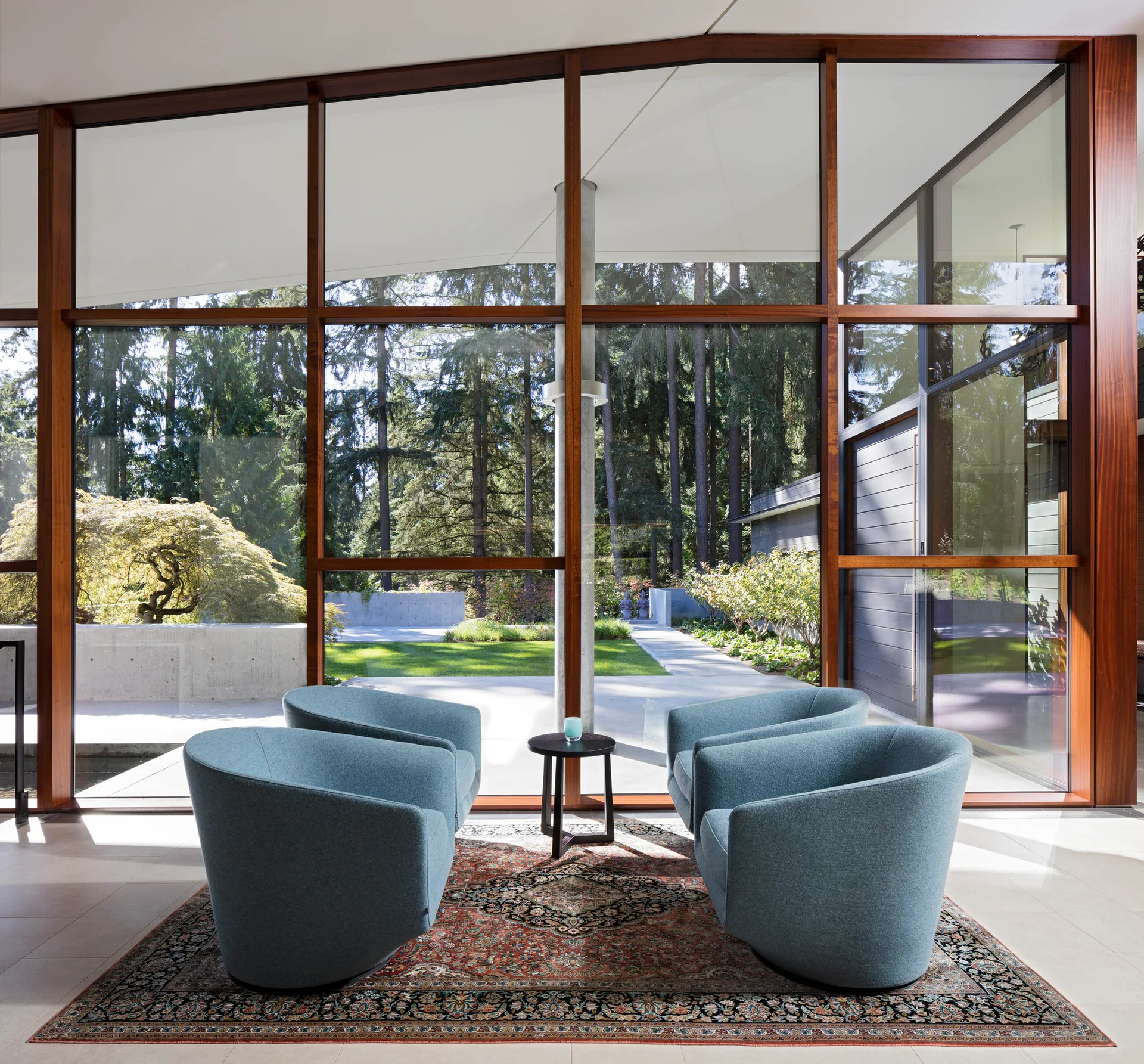 Four blue armchairs sit symmetrically on a patterned traditional rug facing a large floor-to-ceiling glass wall with views of a forest.