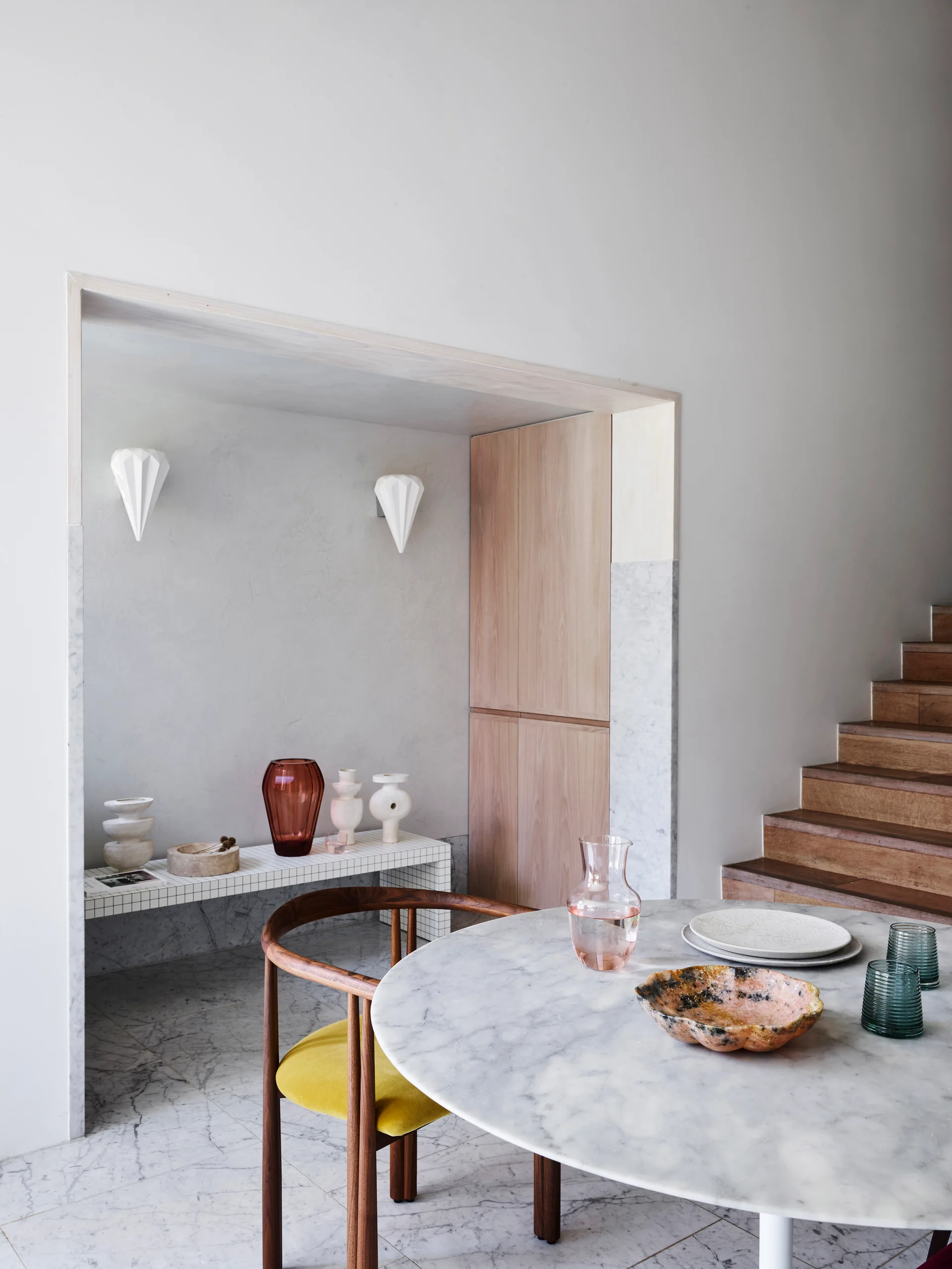 A minimalist dining area features a round marble table, a wood armchair with a yellow cushion, and a tiled alcove displaying decorative vases.