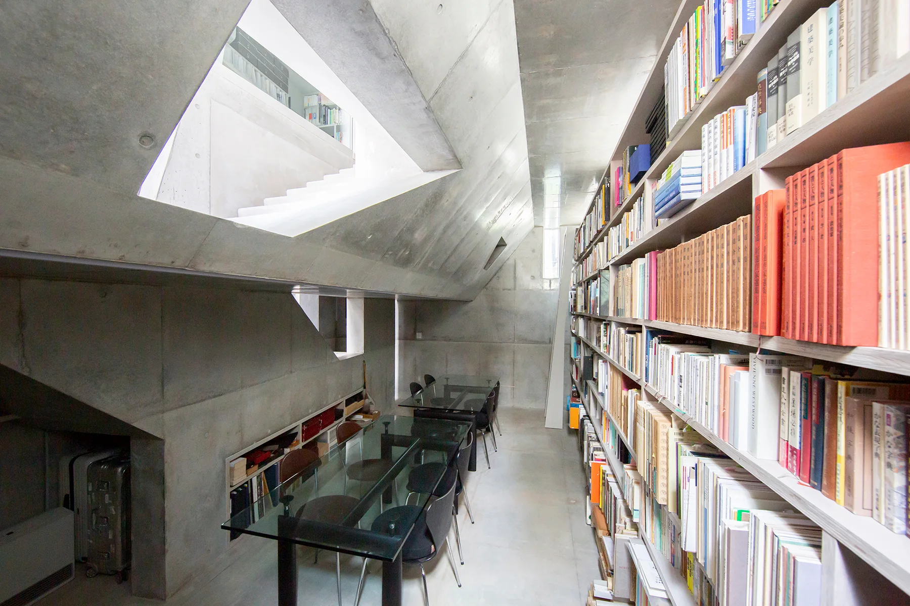 A narrow library featuring exposed concrete walls, a massive wooden bookshelf filled with books, and glass-topped tables paired with black chairs.