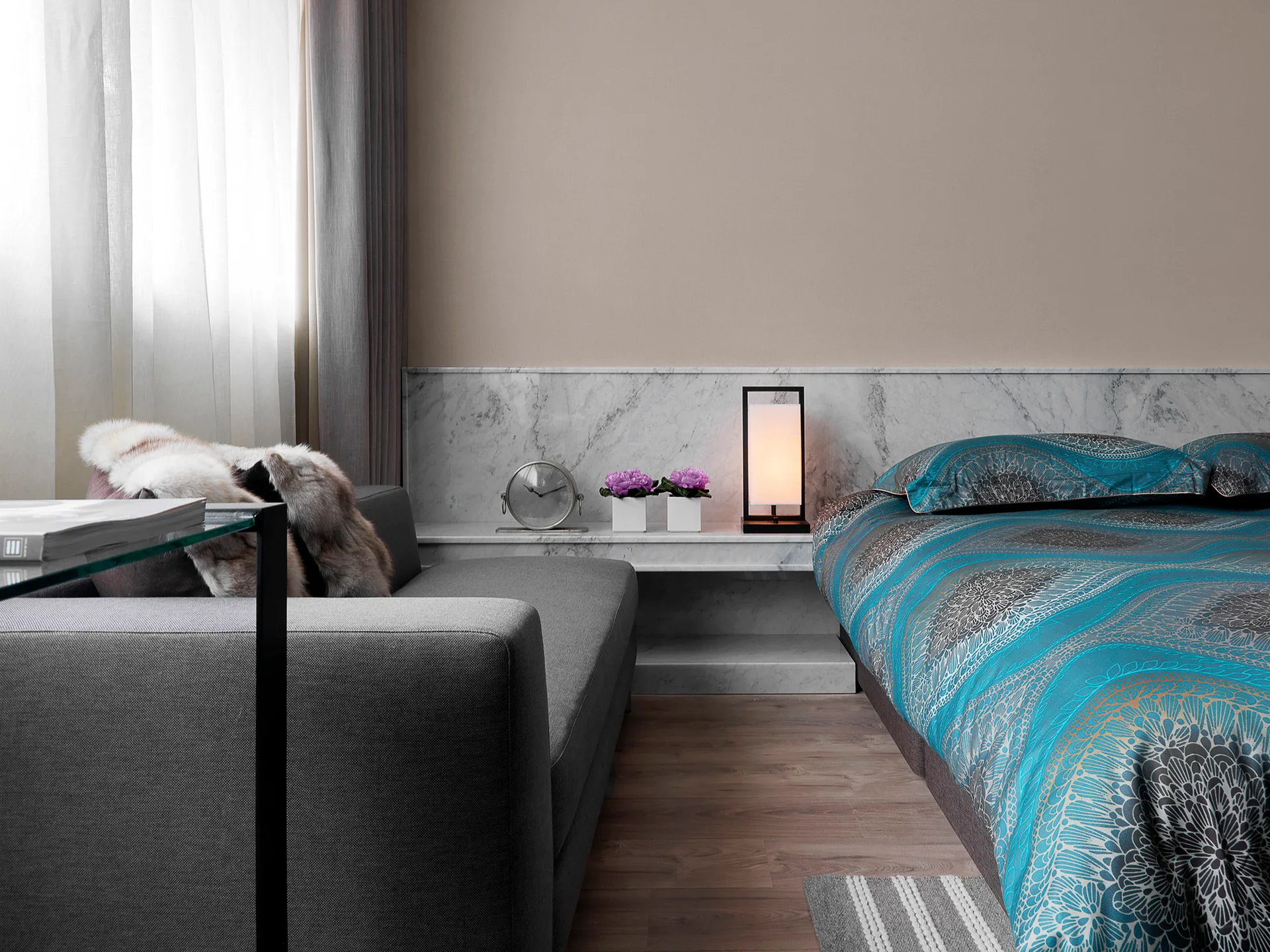A contemporary bedroom featuring a bed with patterned blue bedding next to a grey sofa and an integrated white marble ledge with a glowing lamp.